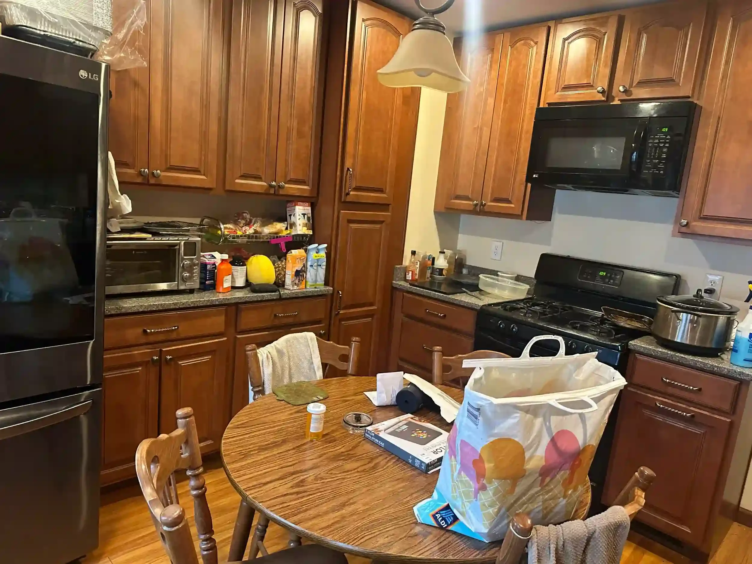 A cluttered kitchen with wooden cabinets, a microwave, and a stove. The kitchen table has various items including a bag of food, a paper towel roll, and some containers. There are chairs around the table, and the kitchen appears to be in use.