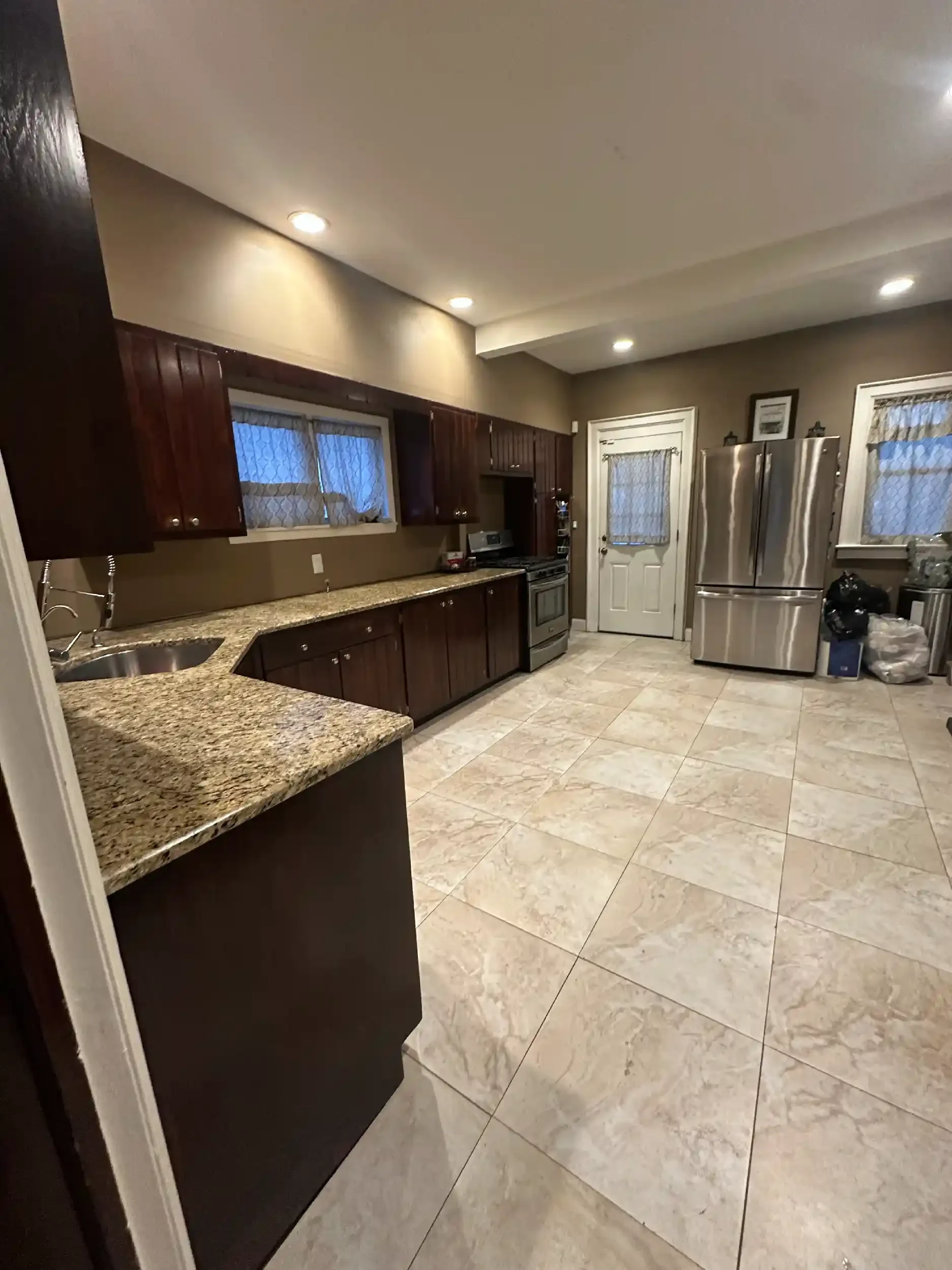 A kitchen with beige tiled floors, dark wood cabinets, granite countertops, a stainless steel refrigerator, and a door with a small window.