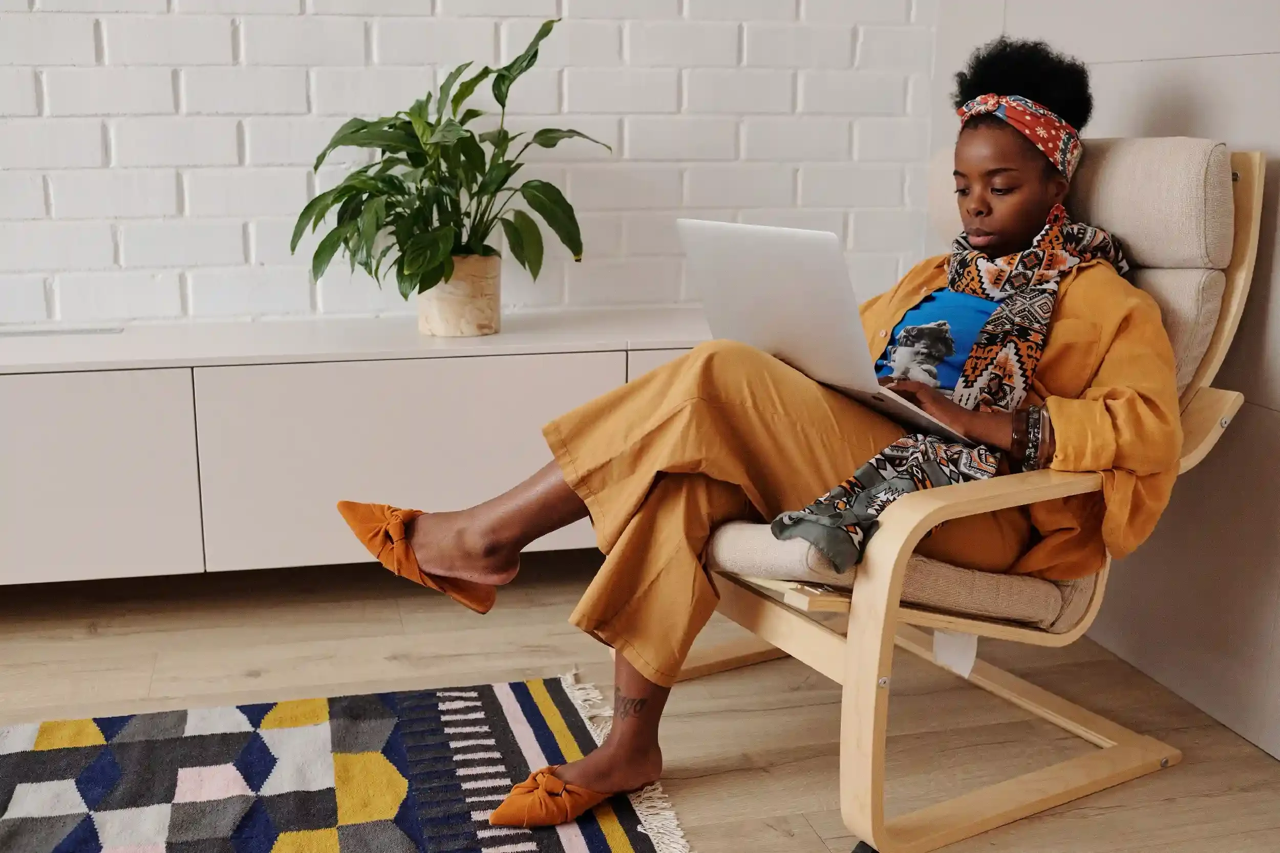 A young woman with dark skin, short curly hair, and a colorful headscarf, sitting in a beige chair against a white brick wall, looking at a laptop. She is dressed in a mustard jacket, a graphic T-shirt, and mustard pants, with orange shoes. There is a white sideboard with a large green potted plant on top and a multicolored geometric rug on the floor.