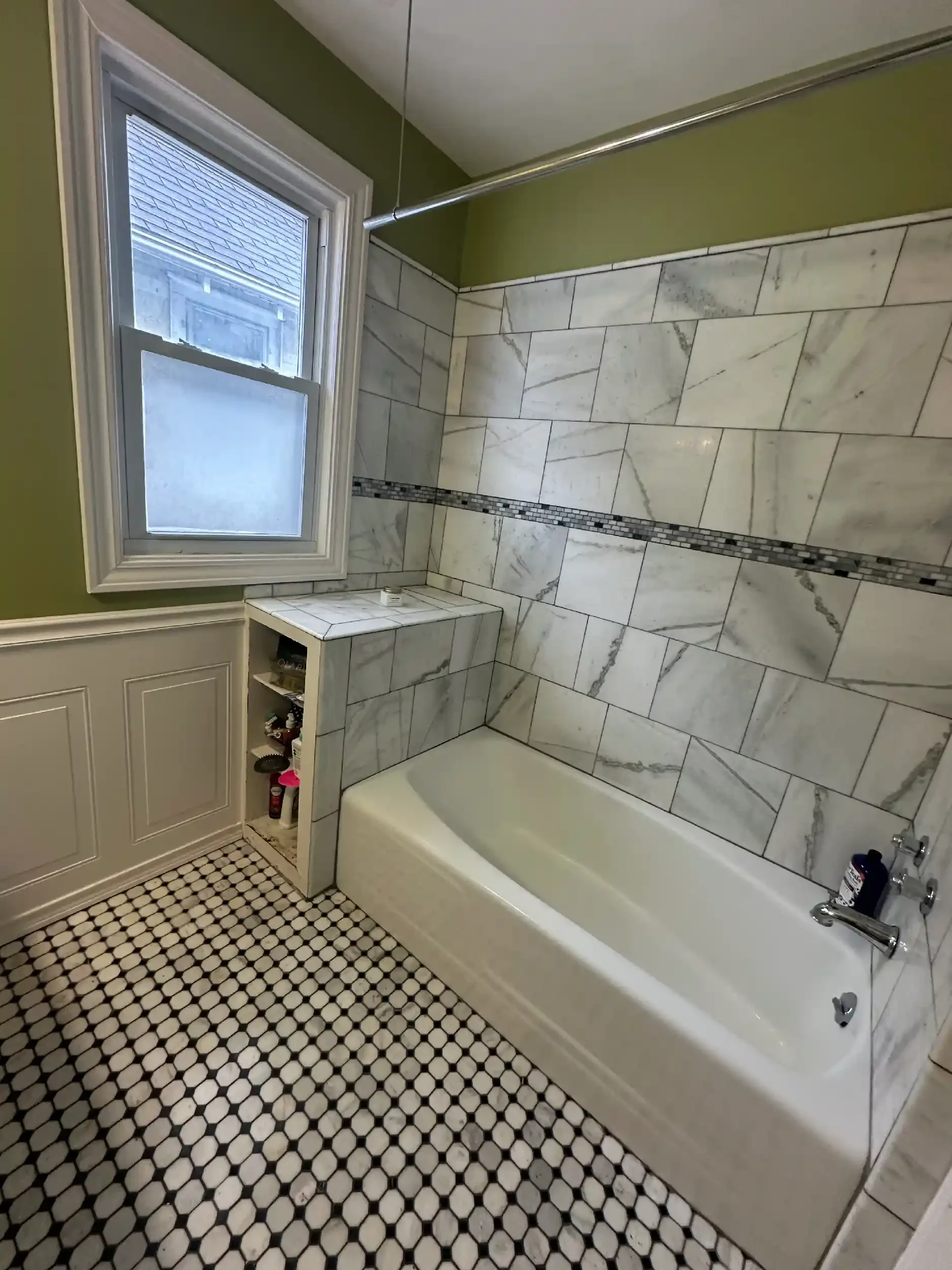 Bathroom with black and white hexagonal tile floor, a bathtub against a wall with marble tiles, a small built-in shelf with toiletries, a window with blinds, and a green wall.
