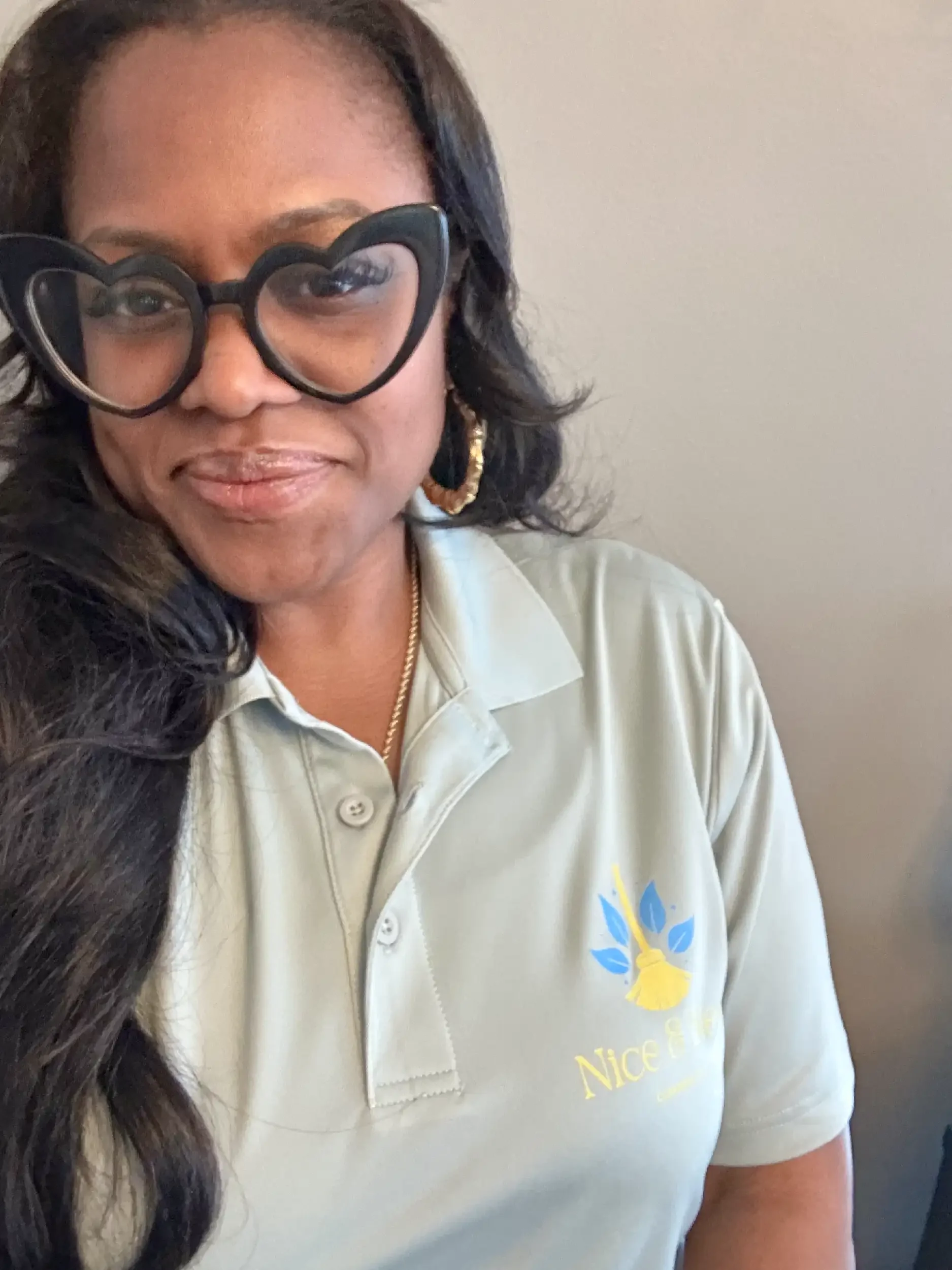 Close-up of a woman wearing large, black, heart-shaped glasses, a light-colored polo shirt with the Nice & Neat Cleaning logo on it, and gold hoop earrings. She has long, dark, wavy hair.