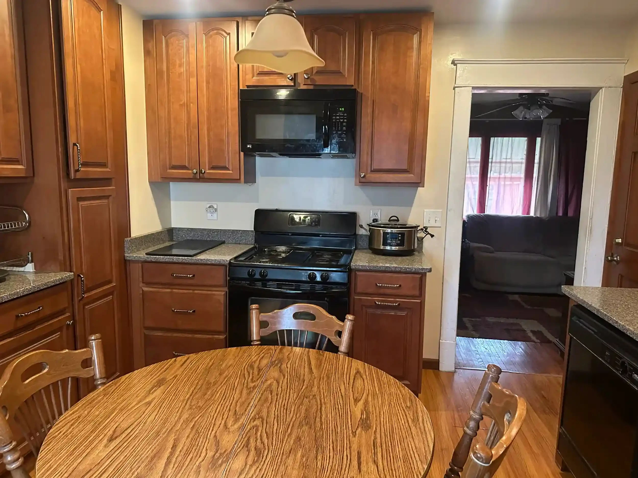 A kitchen with wooden cabinets, a black stove, microwave, and a countertop with a cutting board and a slow cooker. There is a dining table with chairs in the foreground and a doorway leading to a living room with a ceiling fan and large windows.