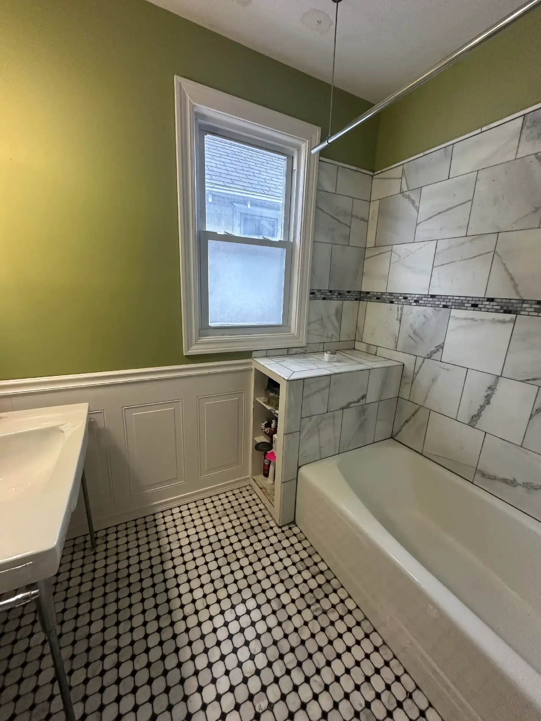 A bathroom with a window, green painted upper wall, white lower wall paneling, marble-tiled shower area with a decorative tile strip, black and white hexagon patterned floor, and a bathtub with a white exterior.