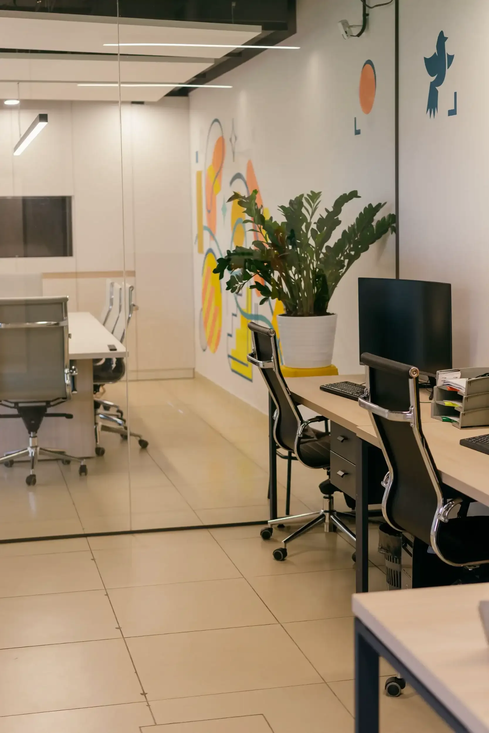 Office space with desks, chairs, computers, and a large plant. The wall is decorated with colorful illustrations of abstract shapes and animals, including a bird and a fish. The room has a modern design with glass partitions.