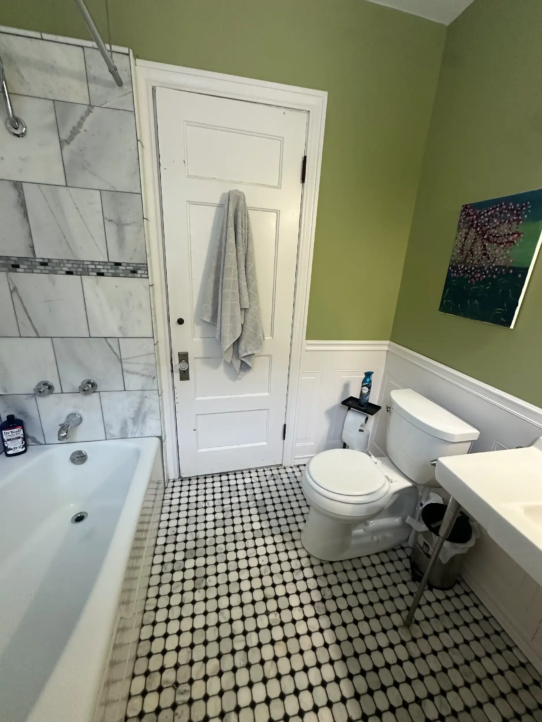 A bathroom with a bathtub on the left, a toilet in the center, and a sink to the right. The wall is painted green, and there is decorative tilework on the floor and walls. A piece of artwork is hanging on the wall.