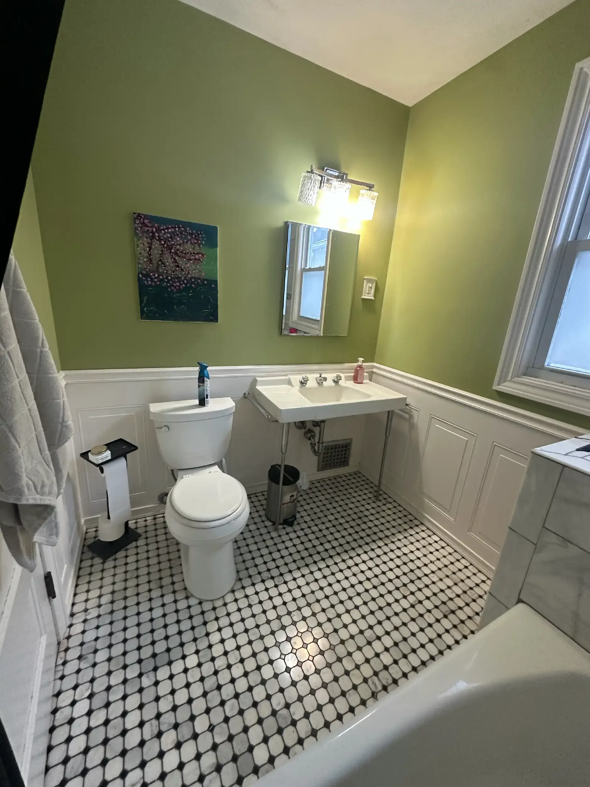 A small bathroom with green walls, a black and white hexagon tile floor, a white sink with chrome fixtures, a mirror, a window, and a white toilet. There is a painting of cherry blossoms on the wall and a light fixture above the mirror.