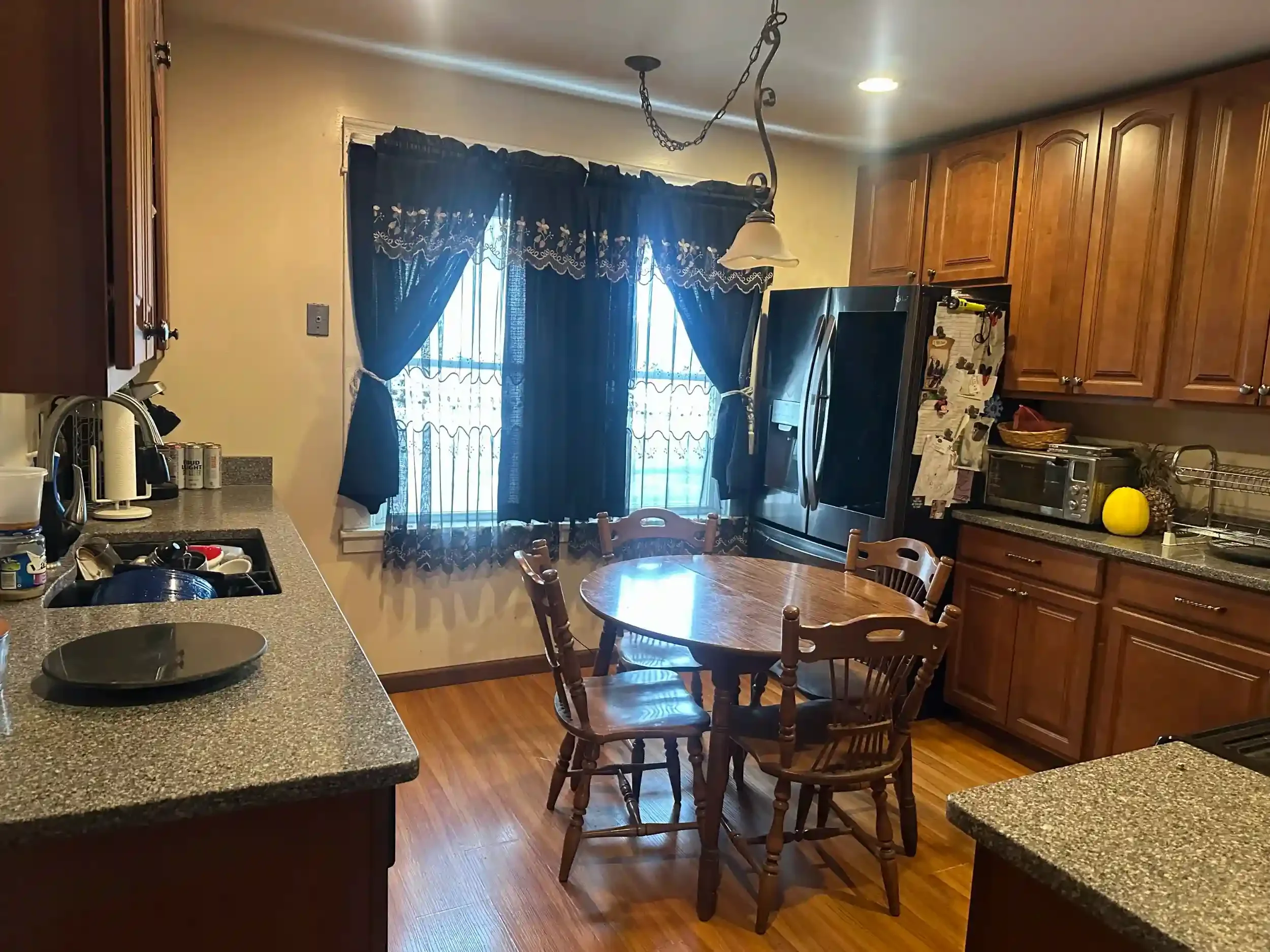 Kitchen with wooden cabinets, a round wooden dining table with chairs, a window with dark blue curtains, and various appliances on the counters.
