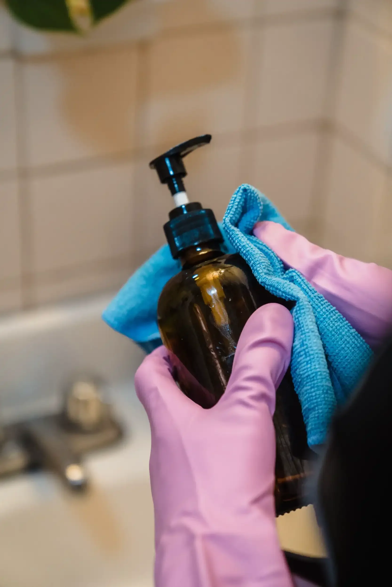 Person wearing pink gloves holding a brown bottle with a pump dispenser and a blue cloth in a bathroom, with a blurred background of tiles and a sink.