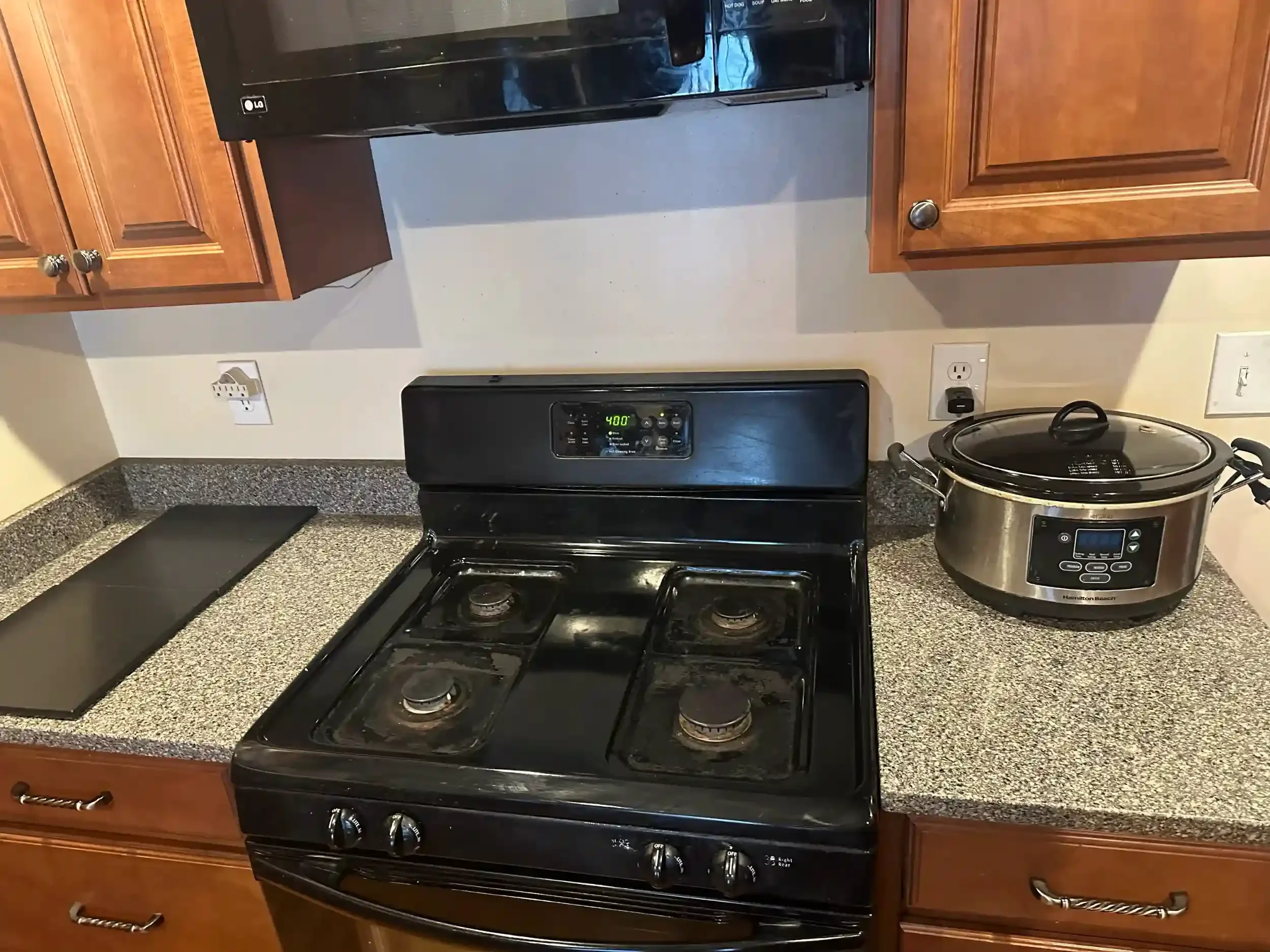Kitchen stove with four burners, a black oven, wooden cabinets, a microwave above the stove, a slow cooker to the right, and electrical outlets on the wall.