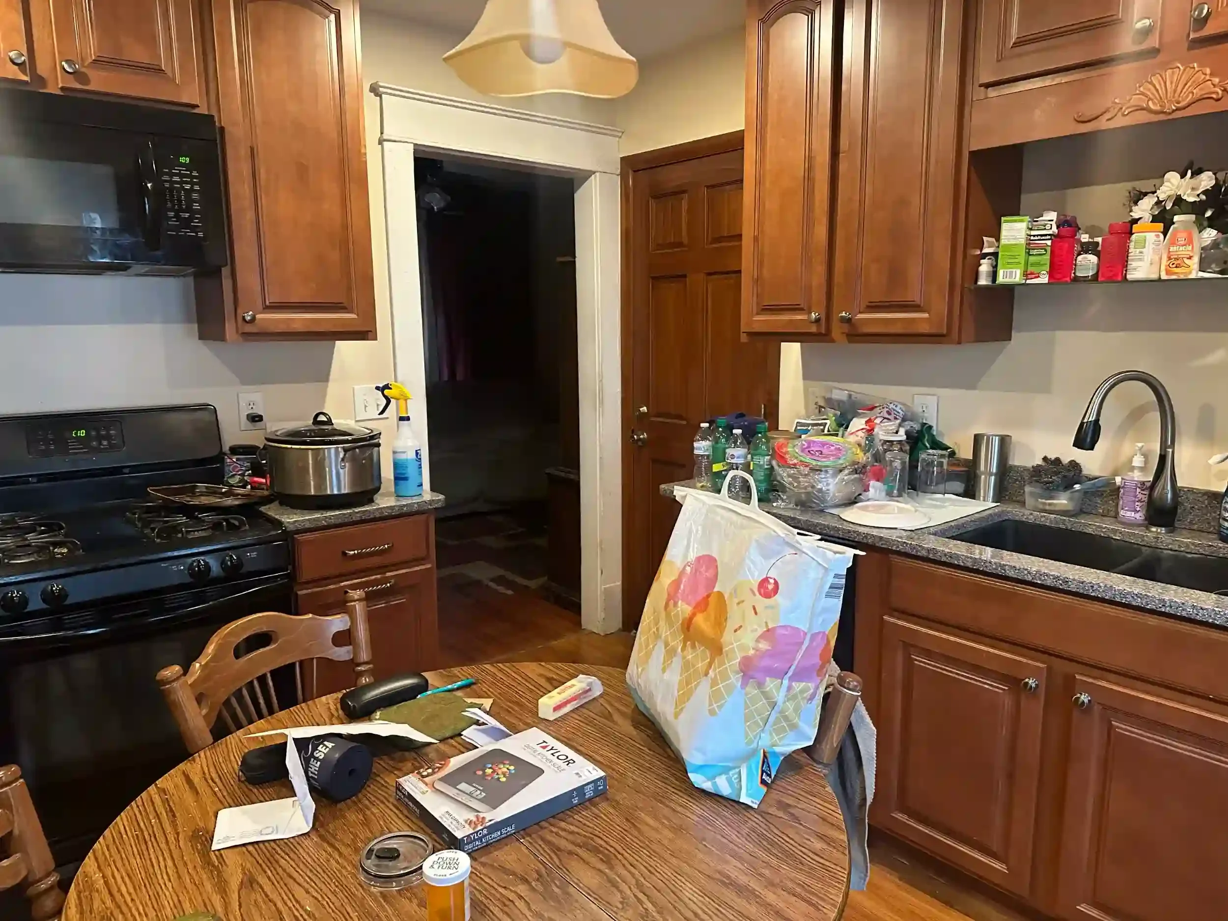 A messy kitchen with cluttered counters and a dining table, wooden cabinets, and appliances.