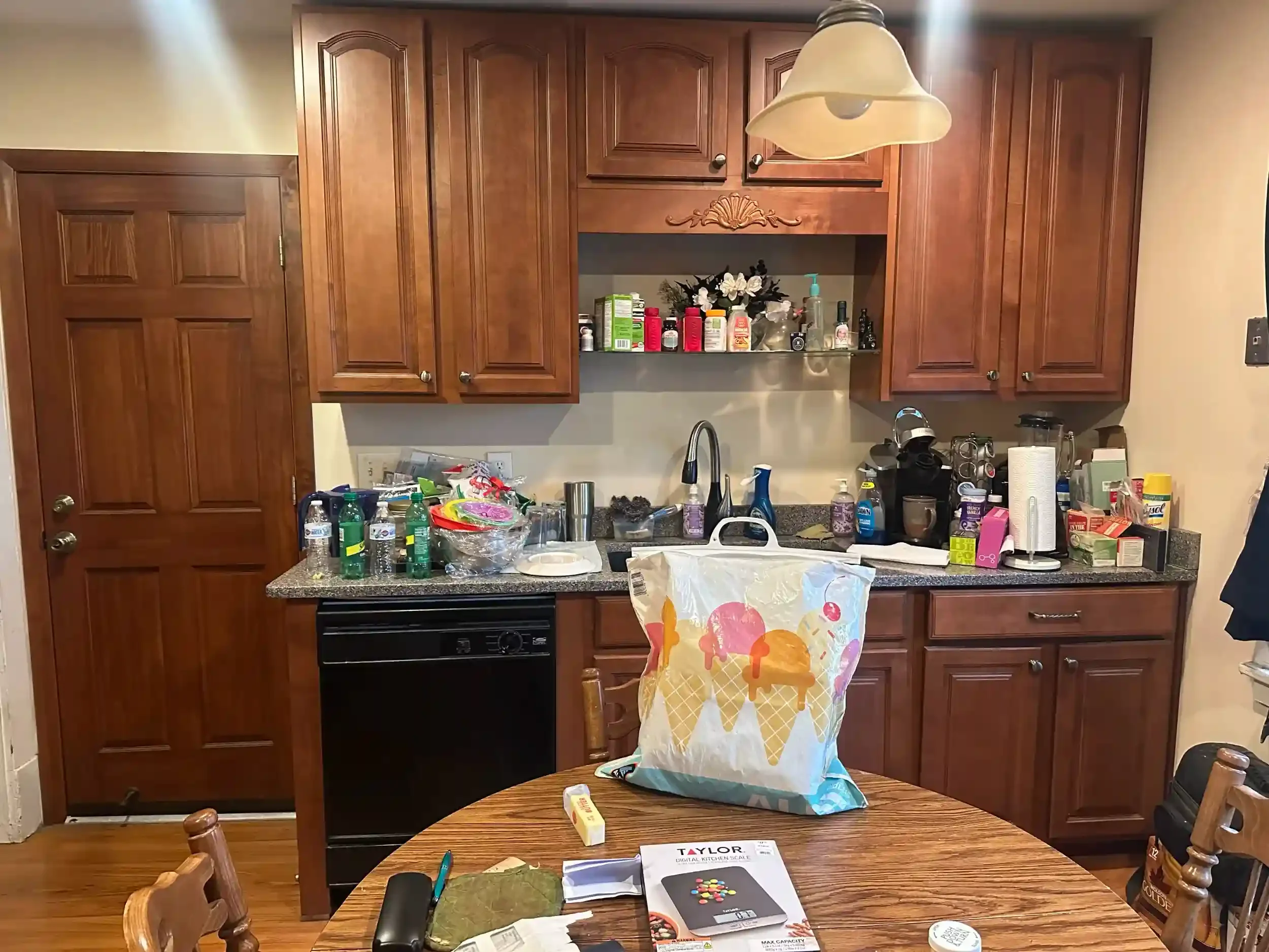 A kitchen with wooden cabinets and a black stove. The countertops are cluttered with various items including water bottles, food packaging, cleaning supplies, and a grocery bag hanging from the counter. There is a table in the foreground with a scale