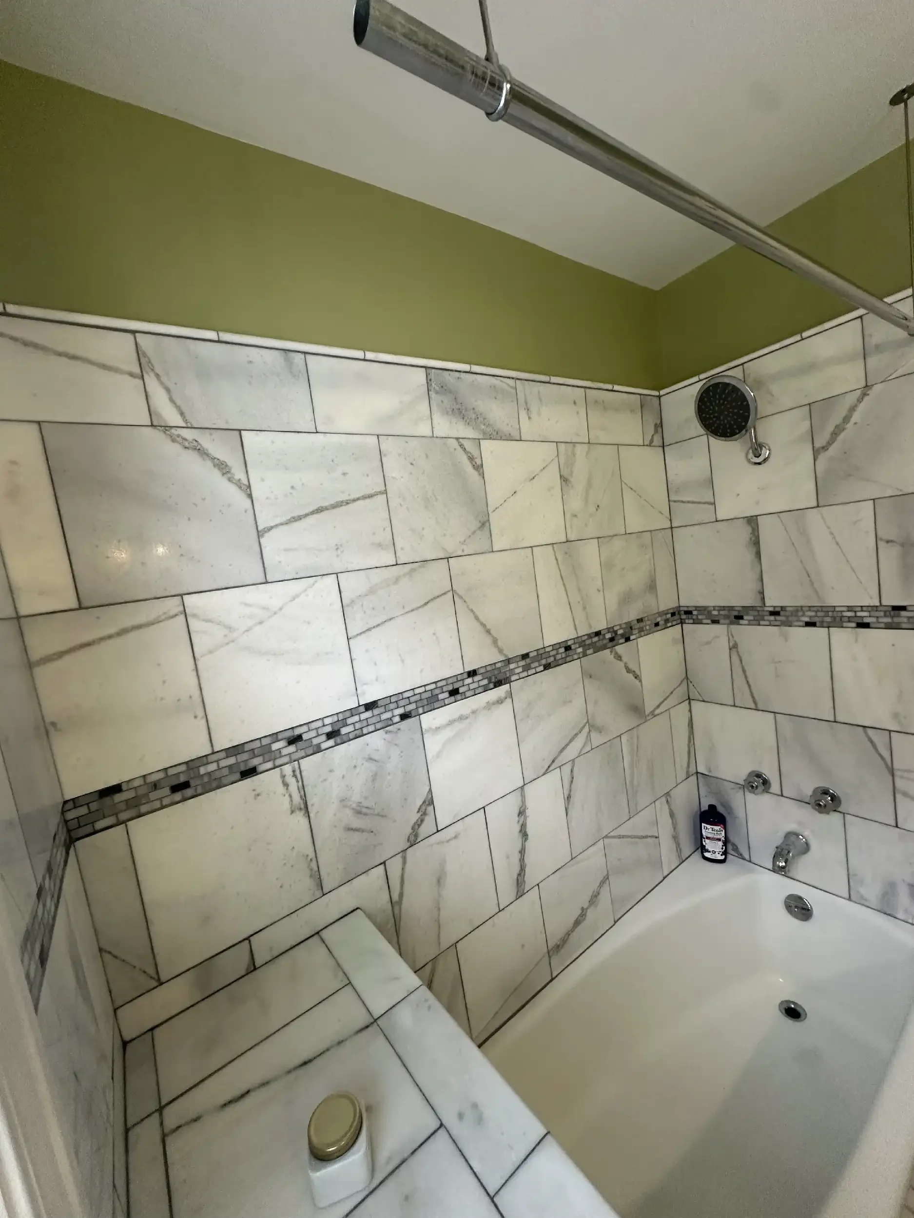 Bathroom with tiled walls and a bathtub, a showerhead, and a bottle of body wash. There is a toilet with a gold-colored lid cover and a small circular ceiling vent.