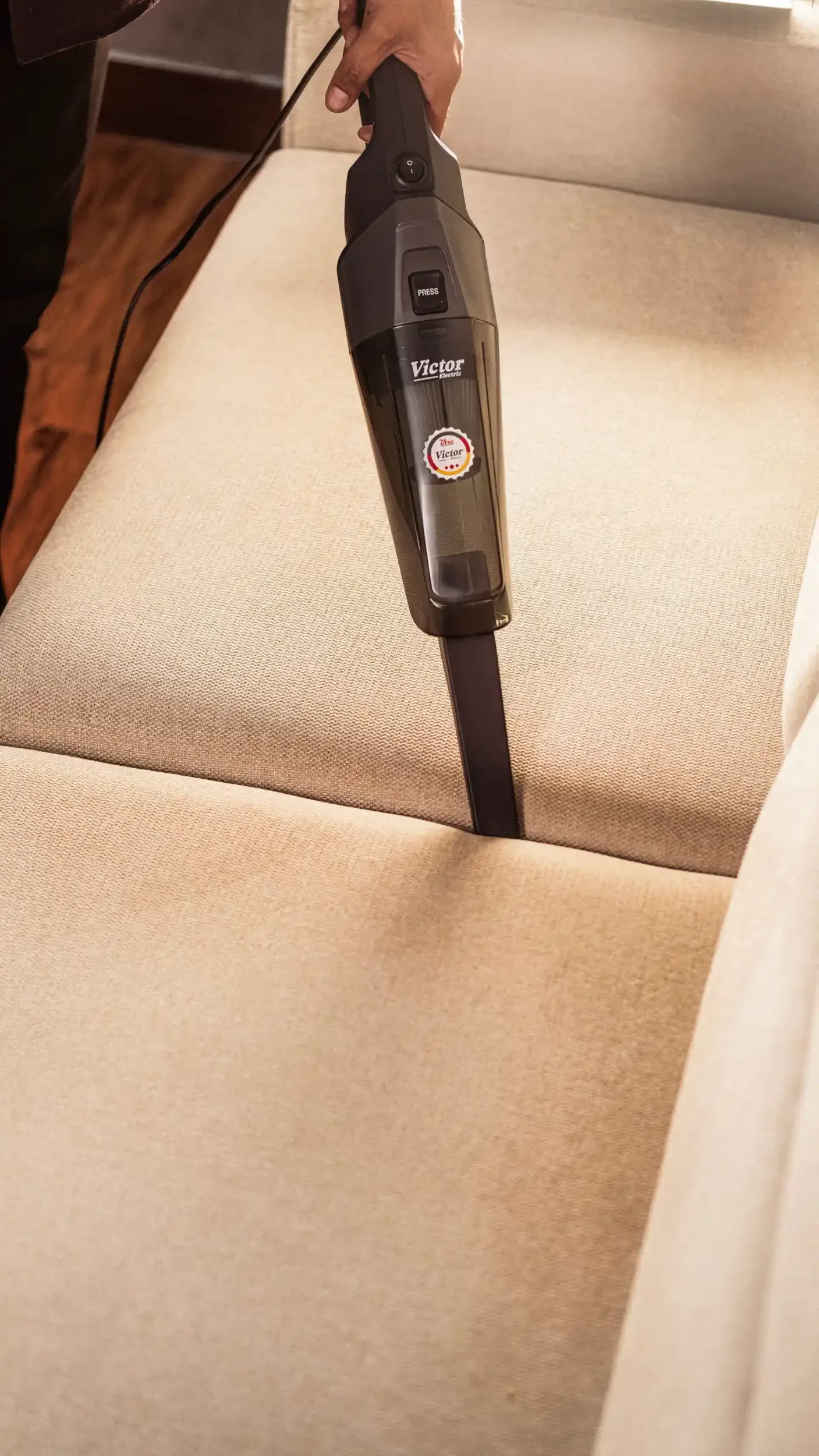 Person using a handheld vacuum cleaner to clean a beige upholstered sofa.
