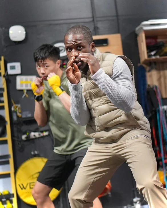 Two men, Coach Shoy in beige workout clothes, and his client, in a green shirt and black shorts, are practicing boxing in a gym. Coach Shoy is in a fighting stance, and the client is slightly behind, mimicking Coach's stance and preparing to box.
