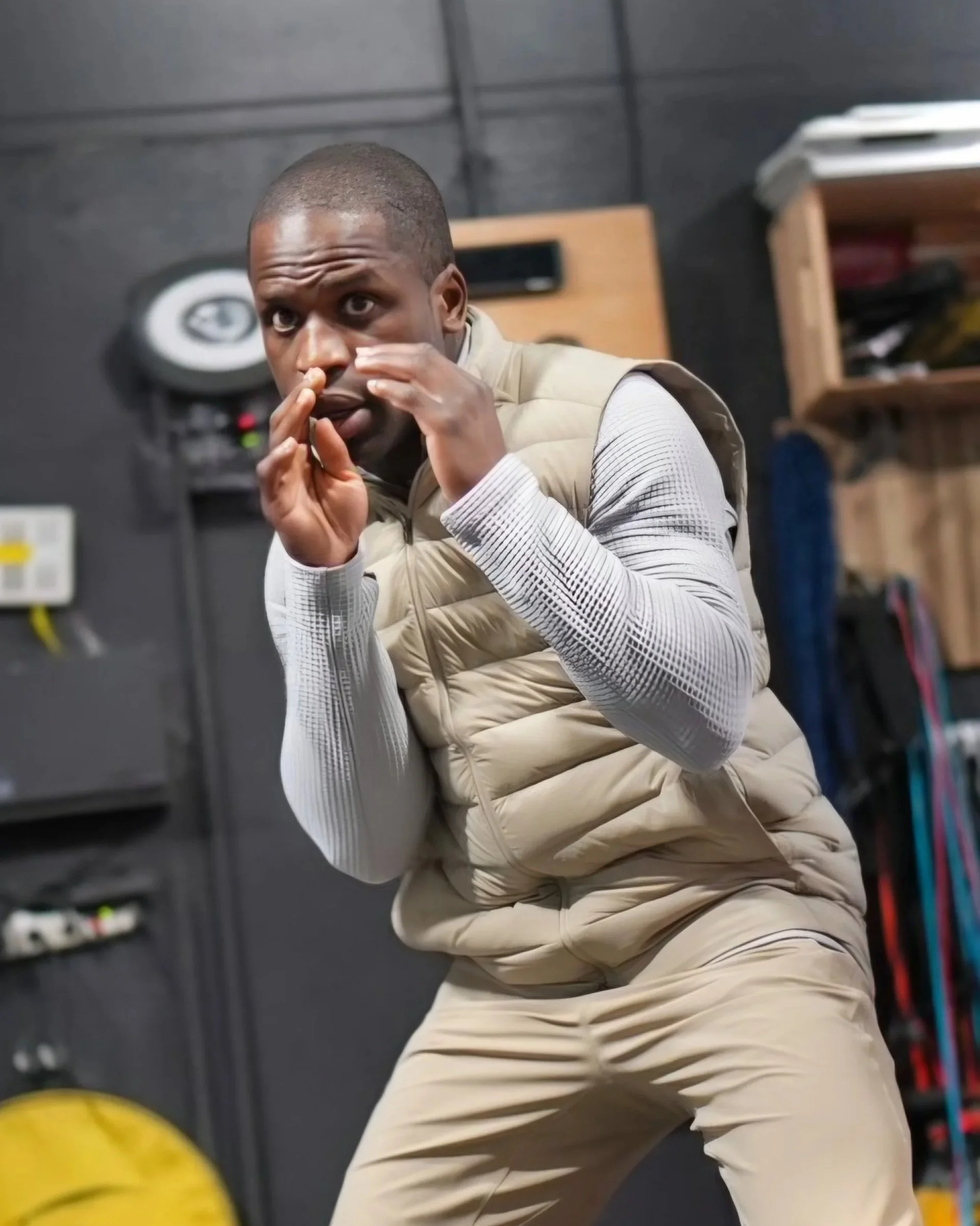 A man in a beige vest and white shirt is preparing to kick a yellow soccer ball indoors. Background includes shelves with papers and equipment.