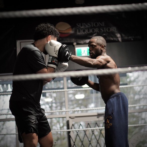 Coach Shoy trains a male client training in a boxing gym, with the client practicing punches while Coach Shoy holds the pads.