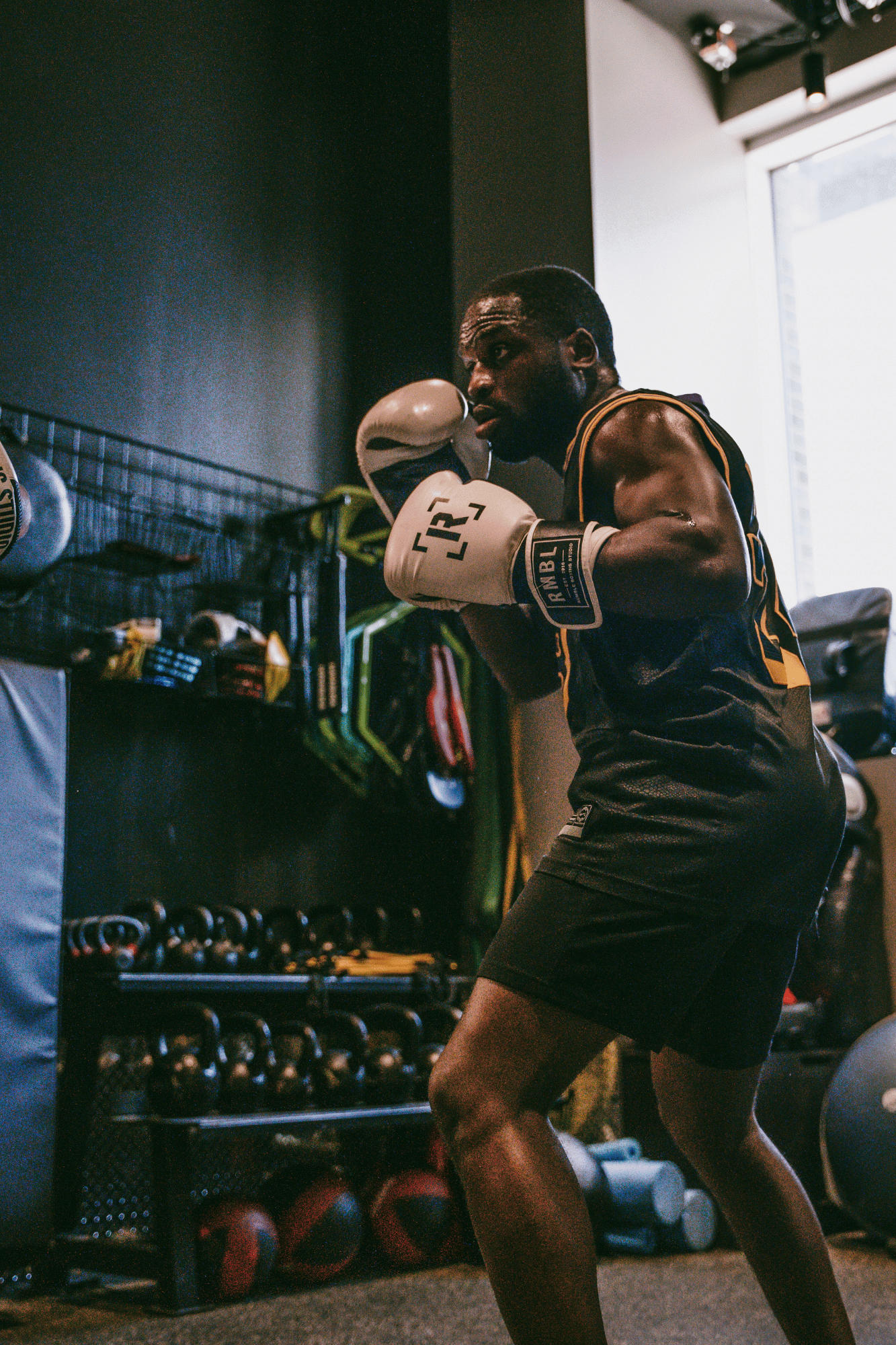 Coach Shoy, a man in boxing gloves practicing punches in a gym, standing in front of a storage rack with boxing gloves and kettlebells, near a window.