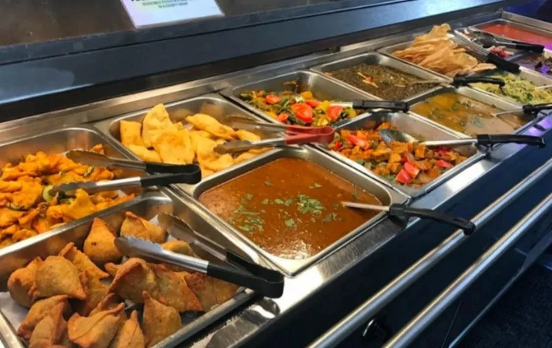 Buffet of various Indian dishes in metal trays, including curry, vegetables, samosas, and fried snacks.