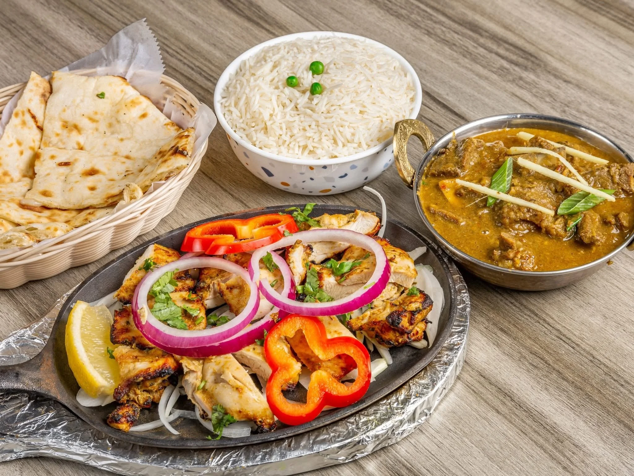 Plate of grilled chicken with sliced red peppers, onions, and lemon, served with rice, naan bread, and a bowl of curry, all on a wooden table.