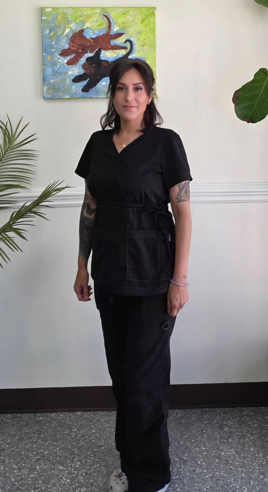 A woman in black scrubs standing inside a room with a white wall, a colorful painting of dogs, and green plants.