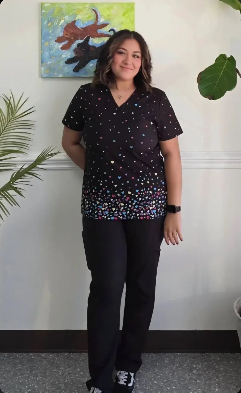 A young woman with shoulder-length wavy brown hair stands indoors in front of a white wall. She is smiling softly, wearing a black shirt with colorful heart patterns and black pants, and has a smartwatch on her left wrist. There are green plants on either side of her and a colorful abstract painting of cats hanging on the wall behind her.