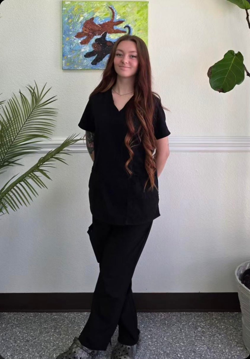 A young woman with long wavy red hair, wearing black scrubs, standing indoors in front of a white wall with a colorful painting of cats and a potted plant with large green leaves.