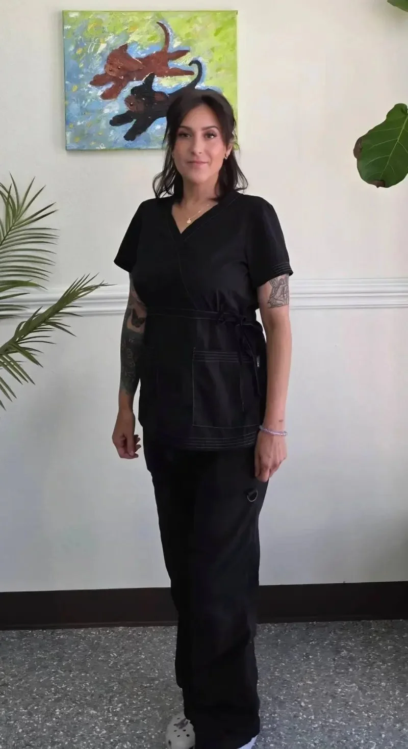 A woman with dark hair wearing a black medical or scrubs uniform standing indoors, with artwork of colorful cats on the wall behind her and plants on either side.
