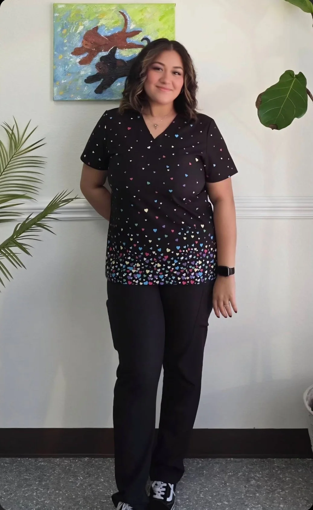 A young woman with shoulder-length wavy hair, wearing a black top decorated with small multicolored hearts and black pants, standing against a white wall. There is a colorful abstract painting of cats behind her and a green plant on the right side.