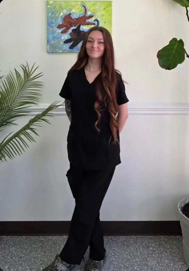 A young woman with long, wavy brown hair and a tattoo on her left arm, wearing black scrubs, standing in front of a white wall with a colorful painting of two black cats, and surrounded by green plants.