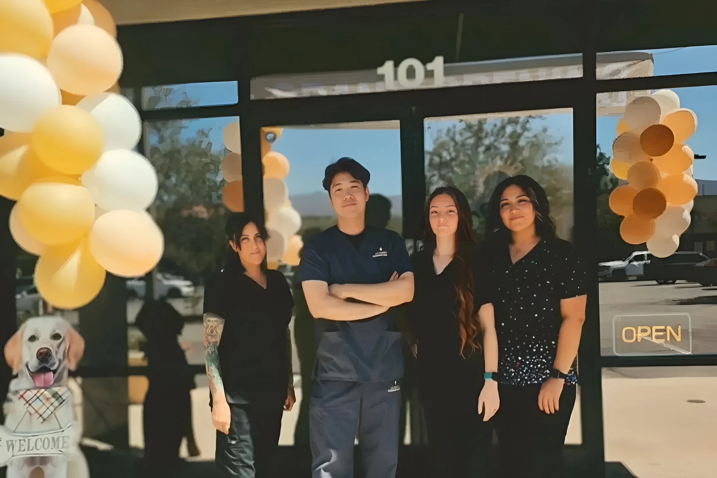 Four people standing in front of a storefront decorated with yellow and white balloons, with a sign indicating the store is open. A dog with care tags is visible in the lower left corner.