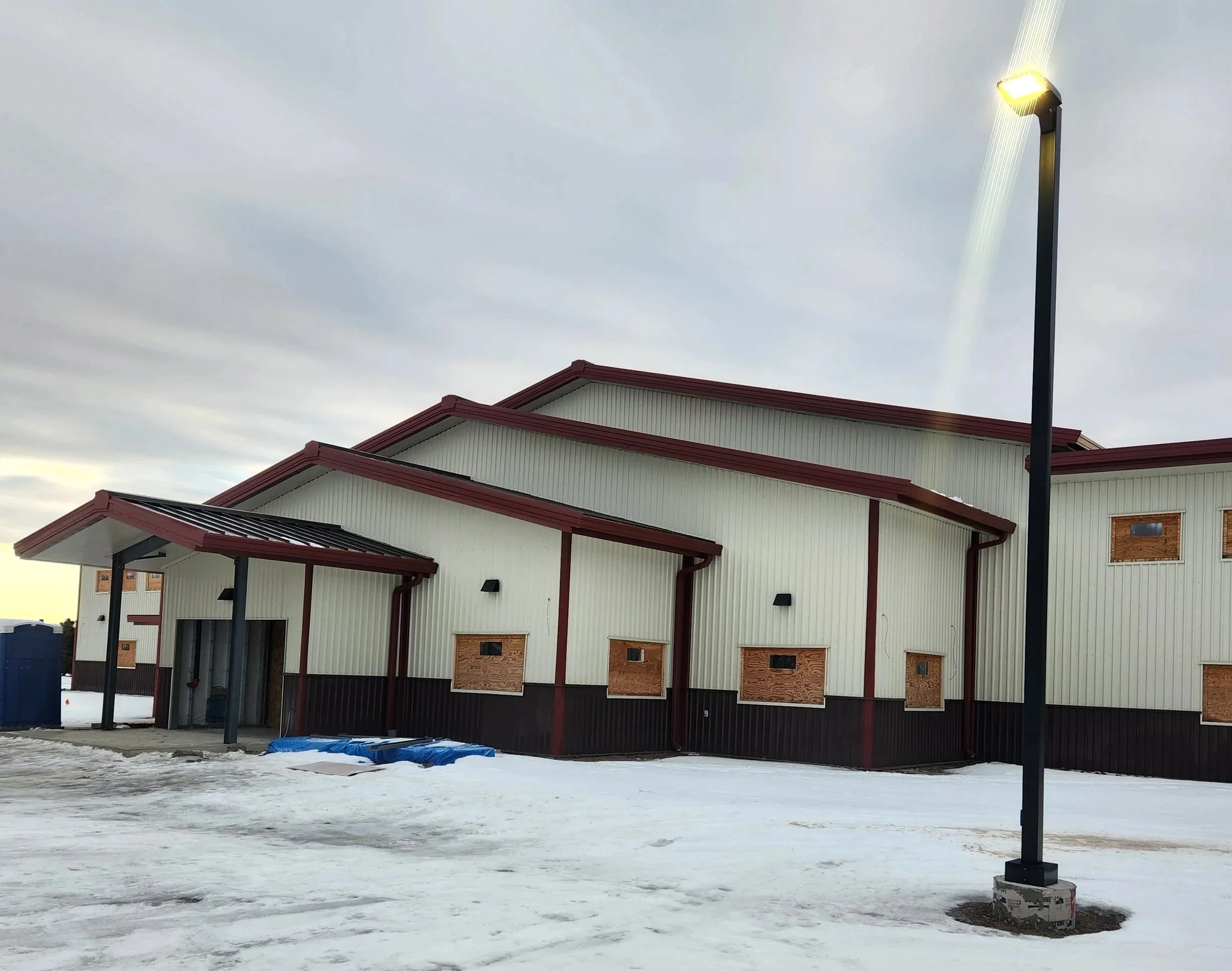 Building under construction with boarded windows, snow on the ground, a light pole, and a cloudy sky.