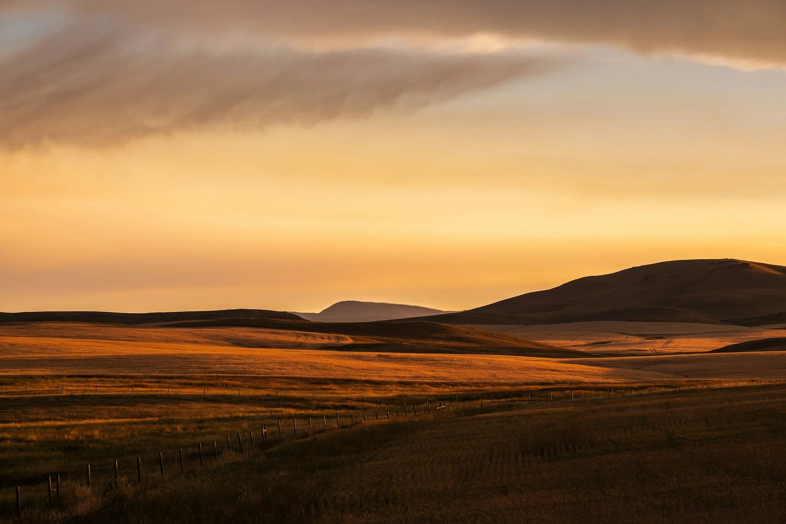 Scenic view of rolling hills and grasslands during sunset, with a partly cloudy sky with orange and yellow hues.