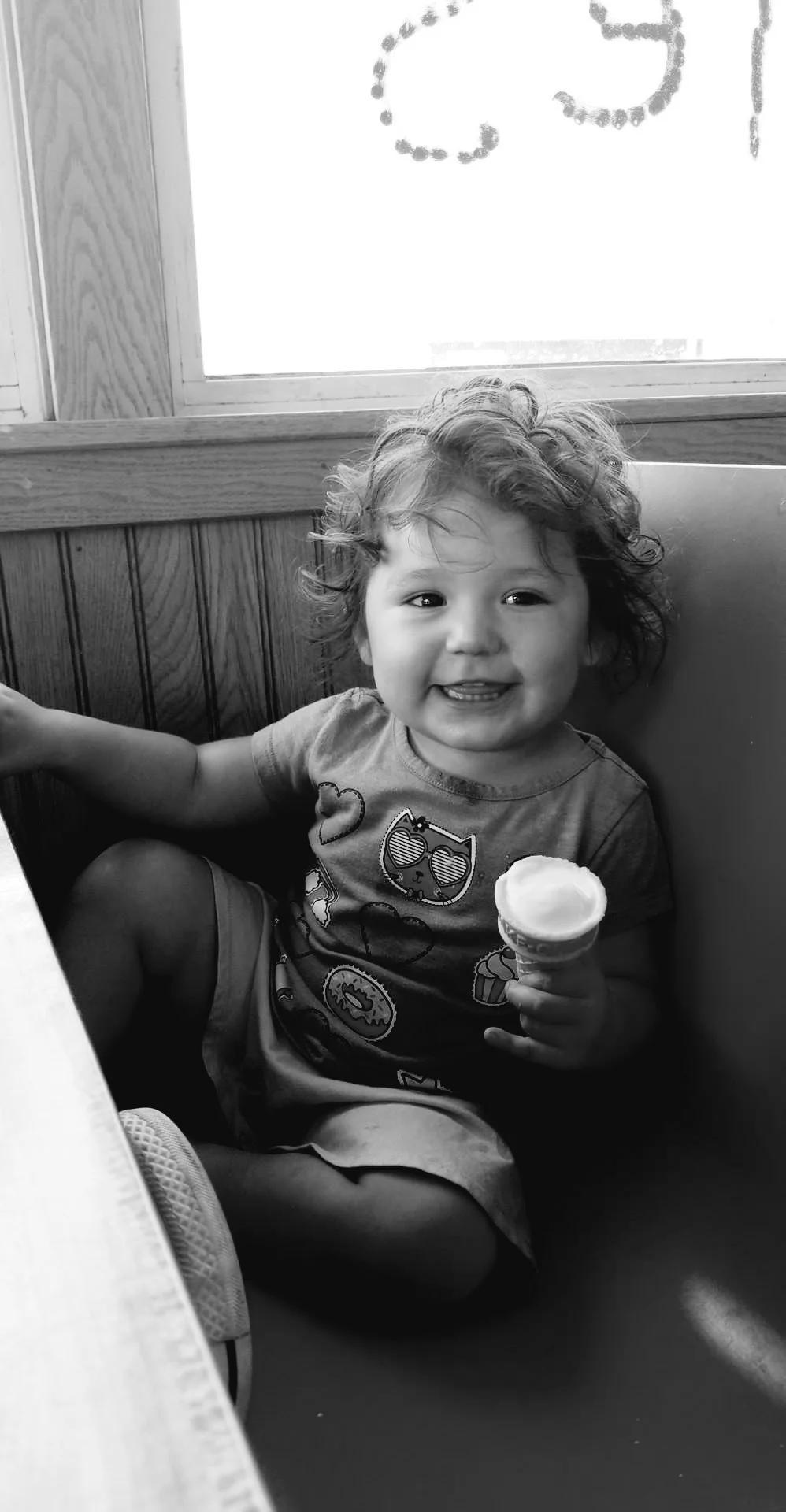 A young child with curly hair sitting in a booth at a restaurant, smiling and holding an ice cream cone. The child is wearing a T-shirt with cartoon animal designs.