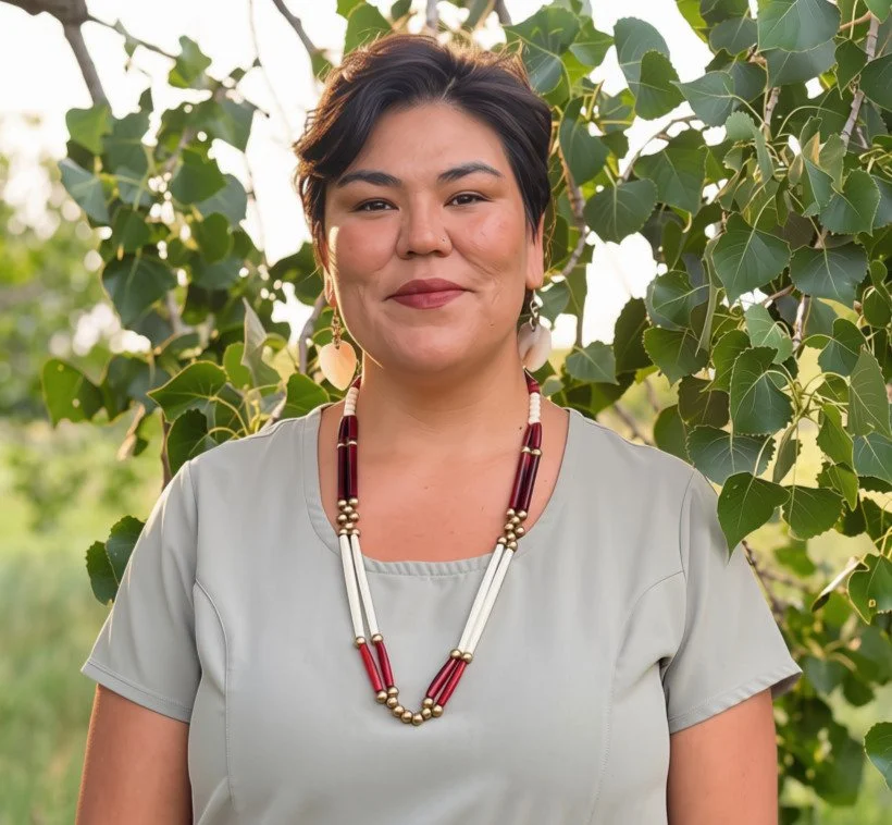 A woman standing outdoors among green leaves, wearing a light-colored top and a red, white, and gold beaded necklace, smiling.