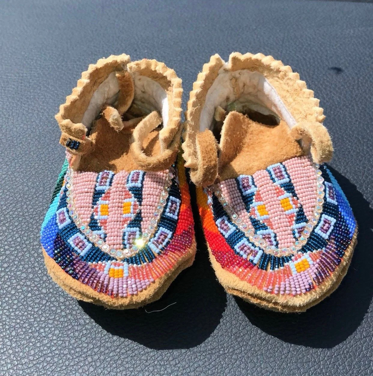 Pair of baby moccasins decorated with colorful beadwork, resting on a dark textured surface.