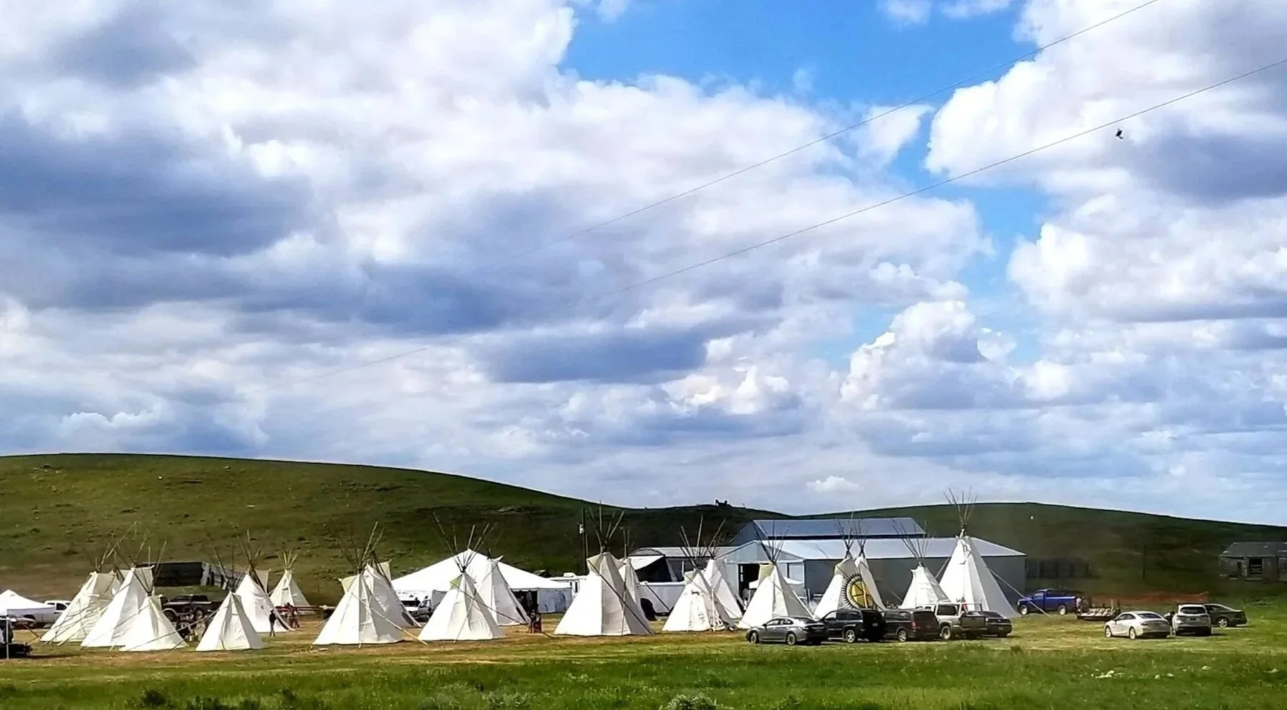 A field with white teepees set up for an event, surrounded by parked cars, with green hills and a partly cloudy sky in the background.