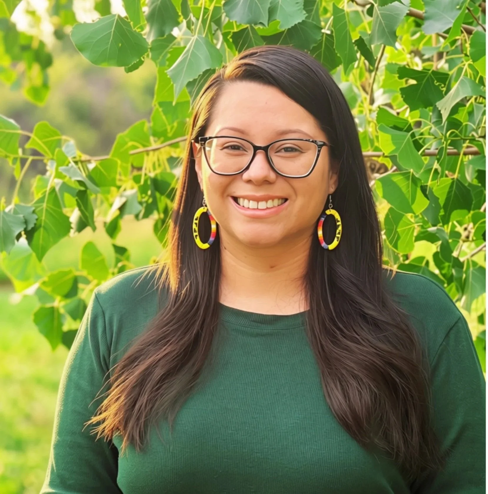 A woman with long dark hair, glasses, and colorful earrings smiles outdoors in front of green leafy plants.