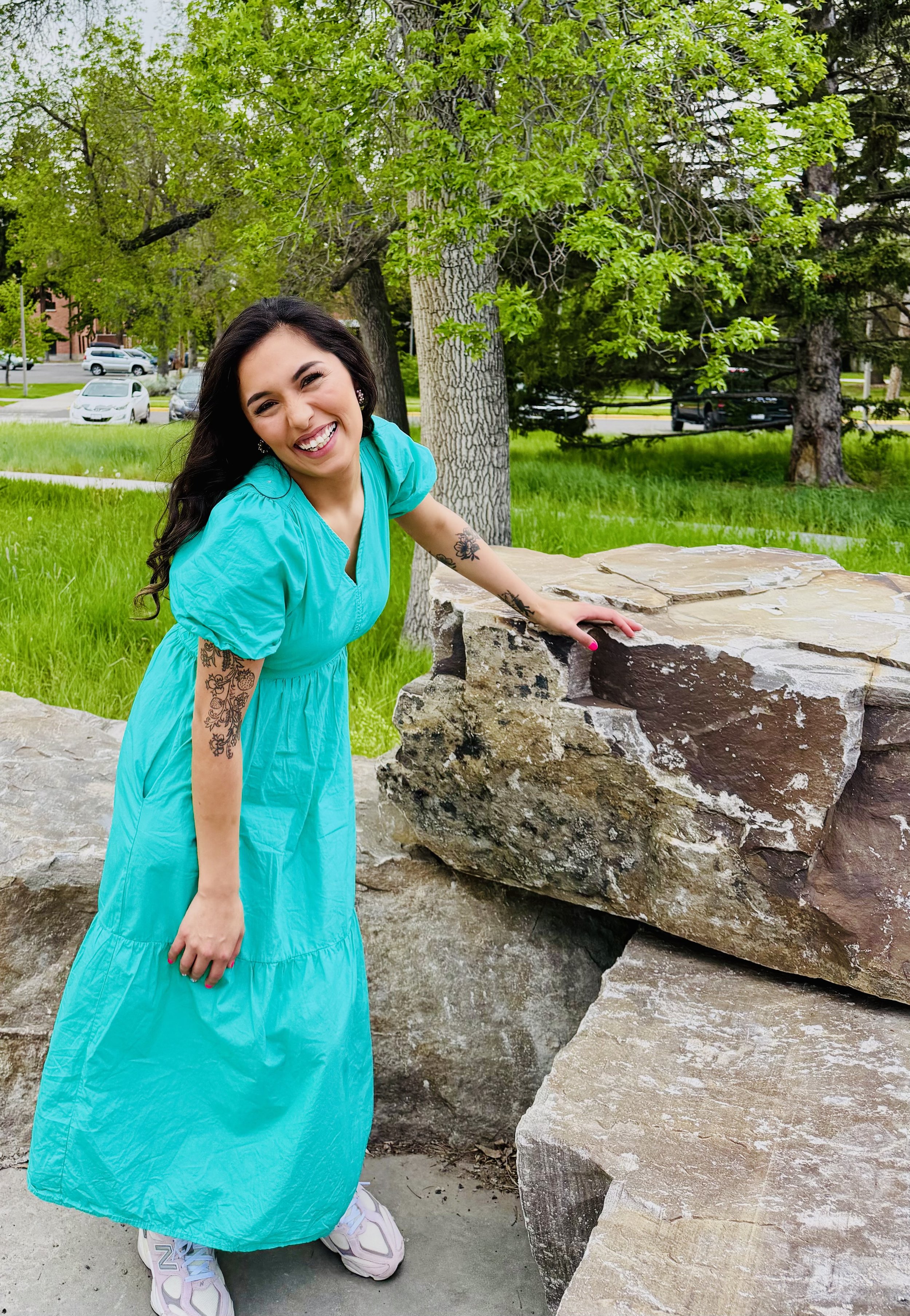 A woman with long dark hair, wearing a bright turquoise dress, smiling and leaning on a large rock in a park with green grass and trees in the background, and cars parked on a street.