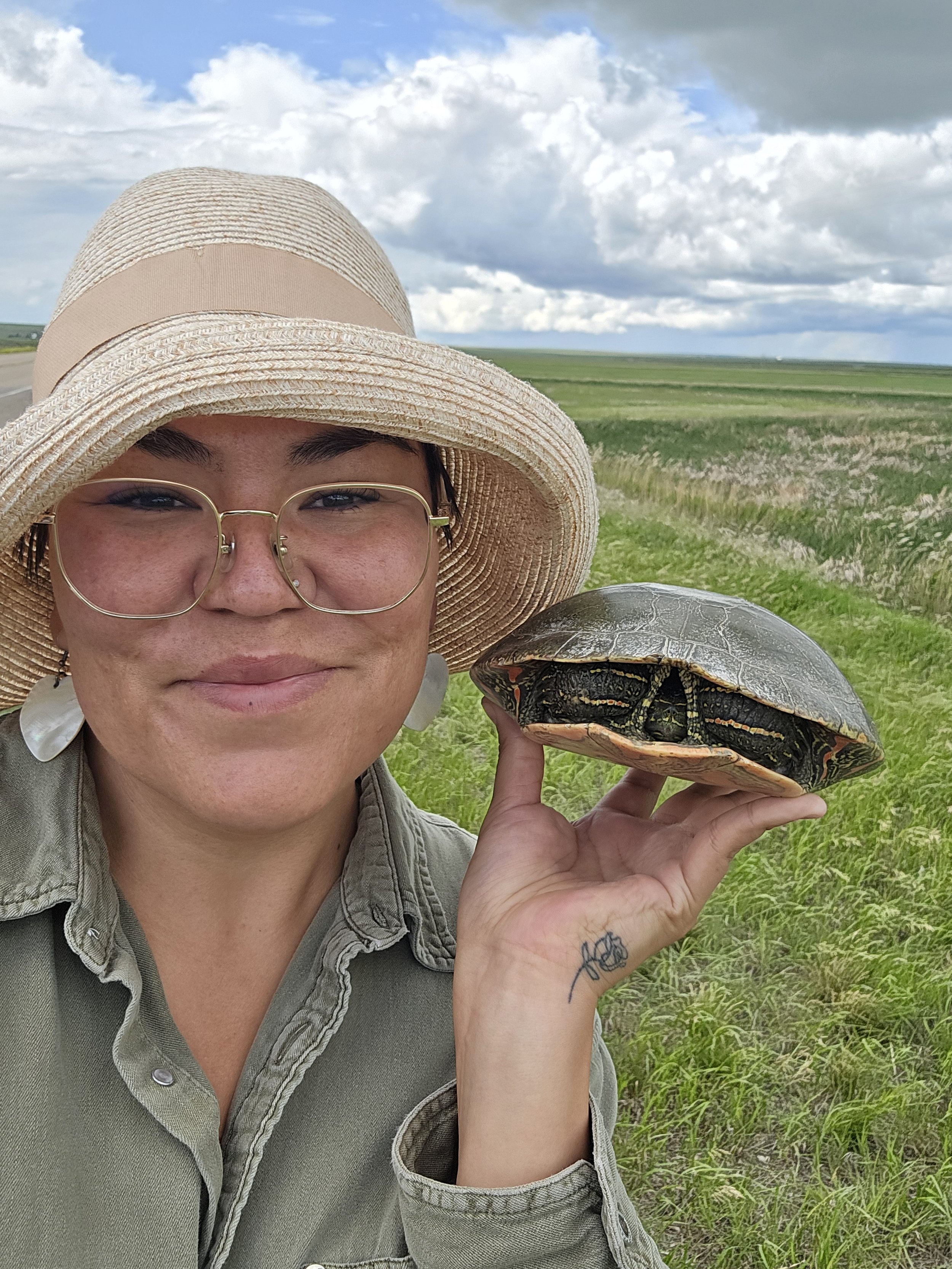 A person wearing a straw hat and glasses holding a turtle with a patterned shell in a grassy field under a cloudy sky.