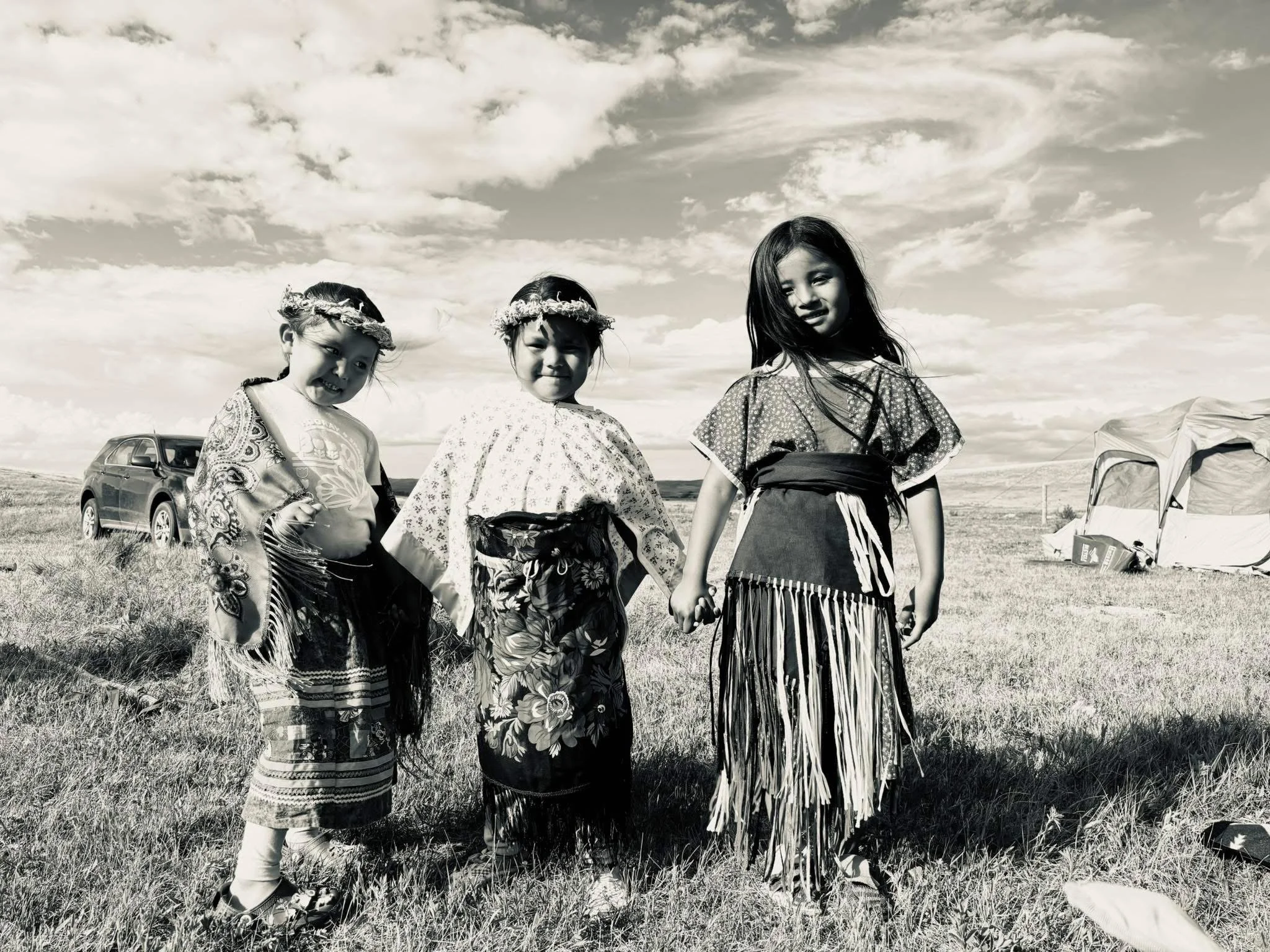 Three young girls dressed in traditional skirts and floral headbands, standing outdoors in a field under a cloudy sky, with a car and tent visible in the background.