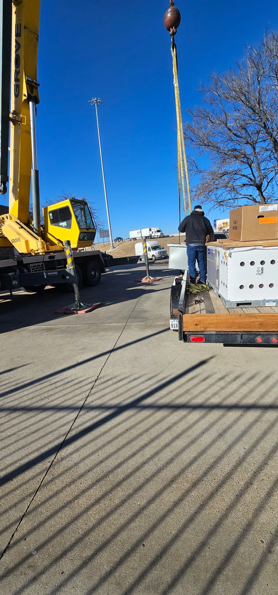 Construction work underway with a crane lifting a large HVAC unit in a mostly clear sky, a worker standing on a trailer, and vehicles on the road in the background.