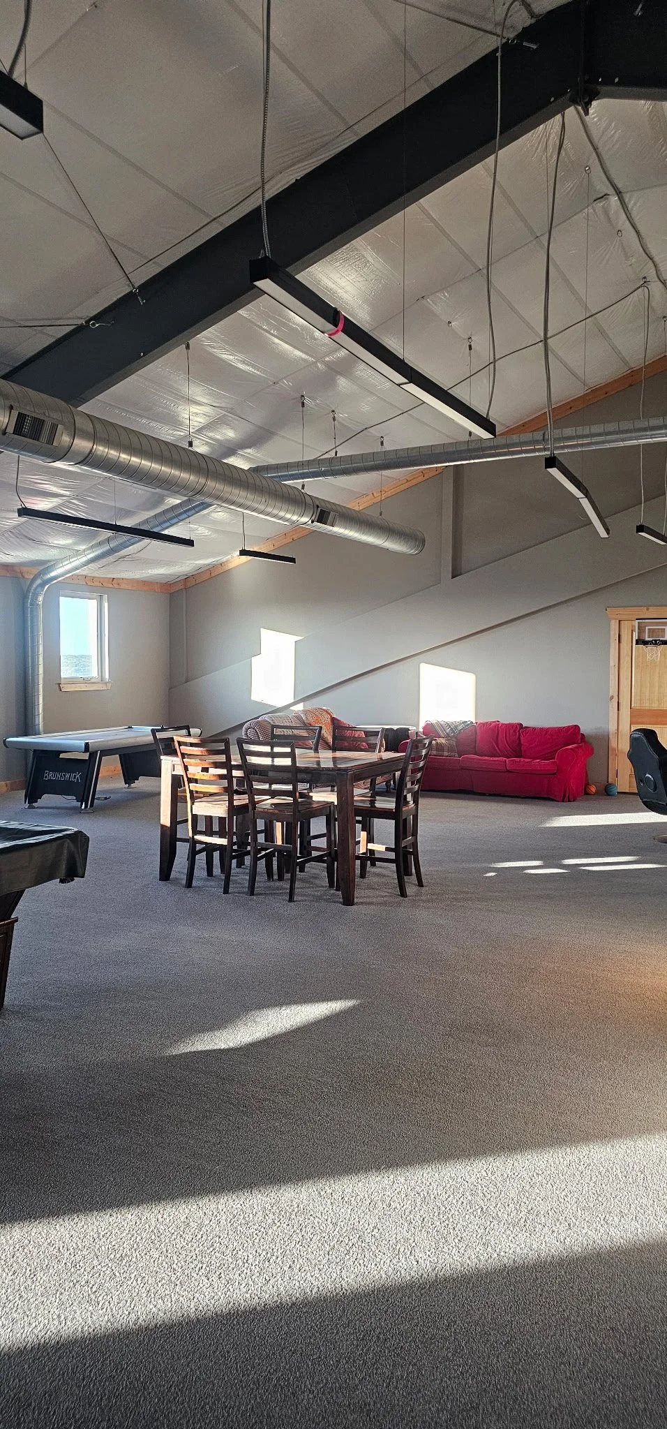 A spacious room with a high ceiling, featuring exposed ductwork and hanging lighting fixtures. There is a red couch, a wooden dining table with chairs, an air hockey table, and some windows letting in natural light.