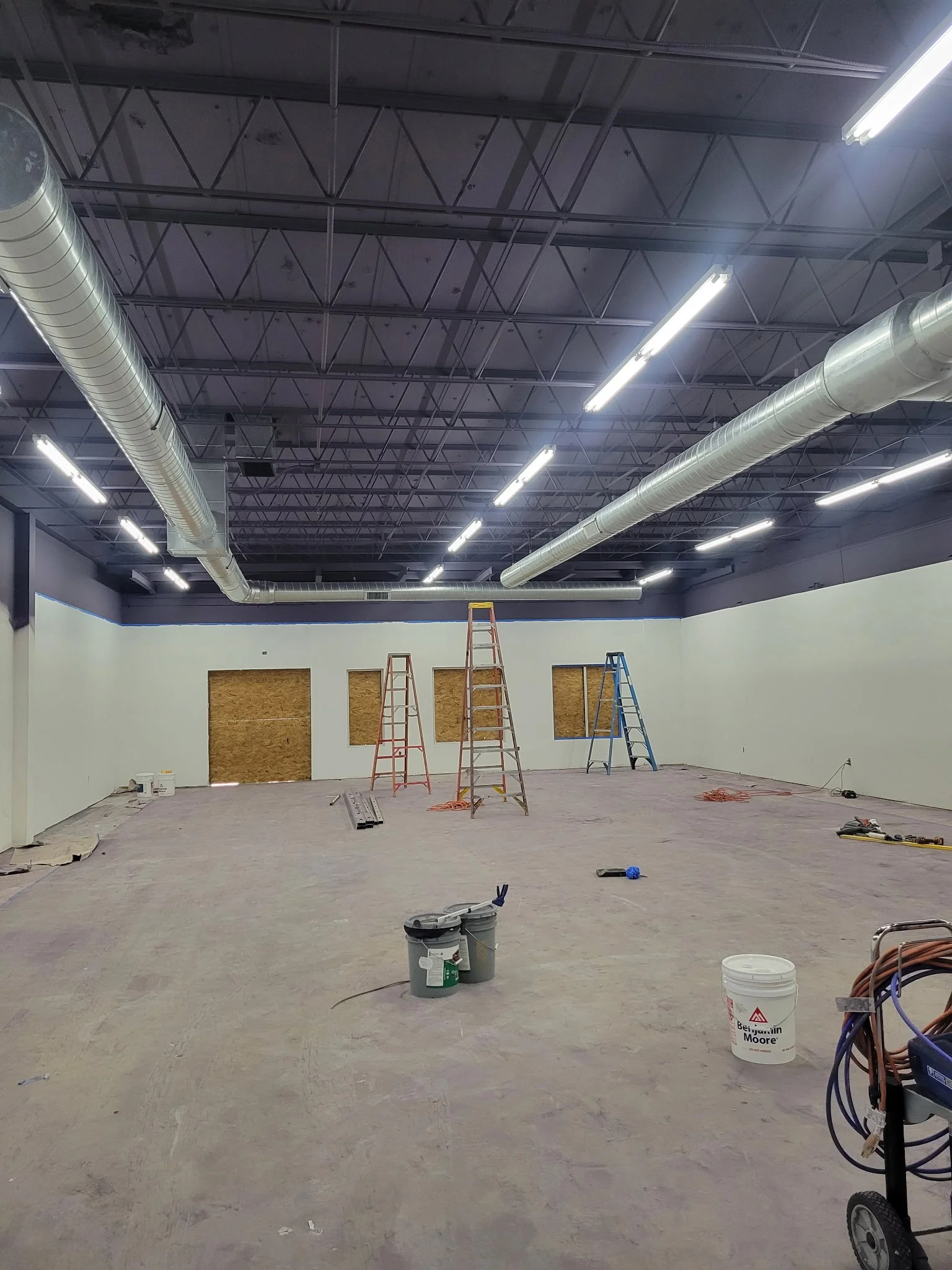 Interior of a building under construction with scaffolding, ladders, and construction materials scattered on the unfinished floor. Exposed ductwork is visible overhead.
