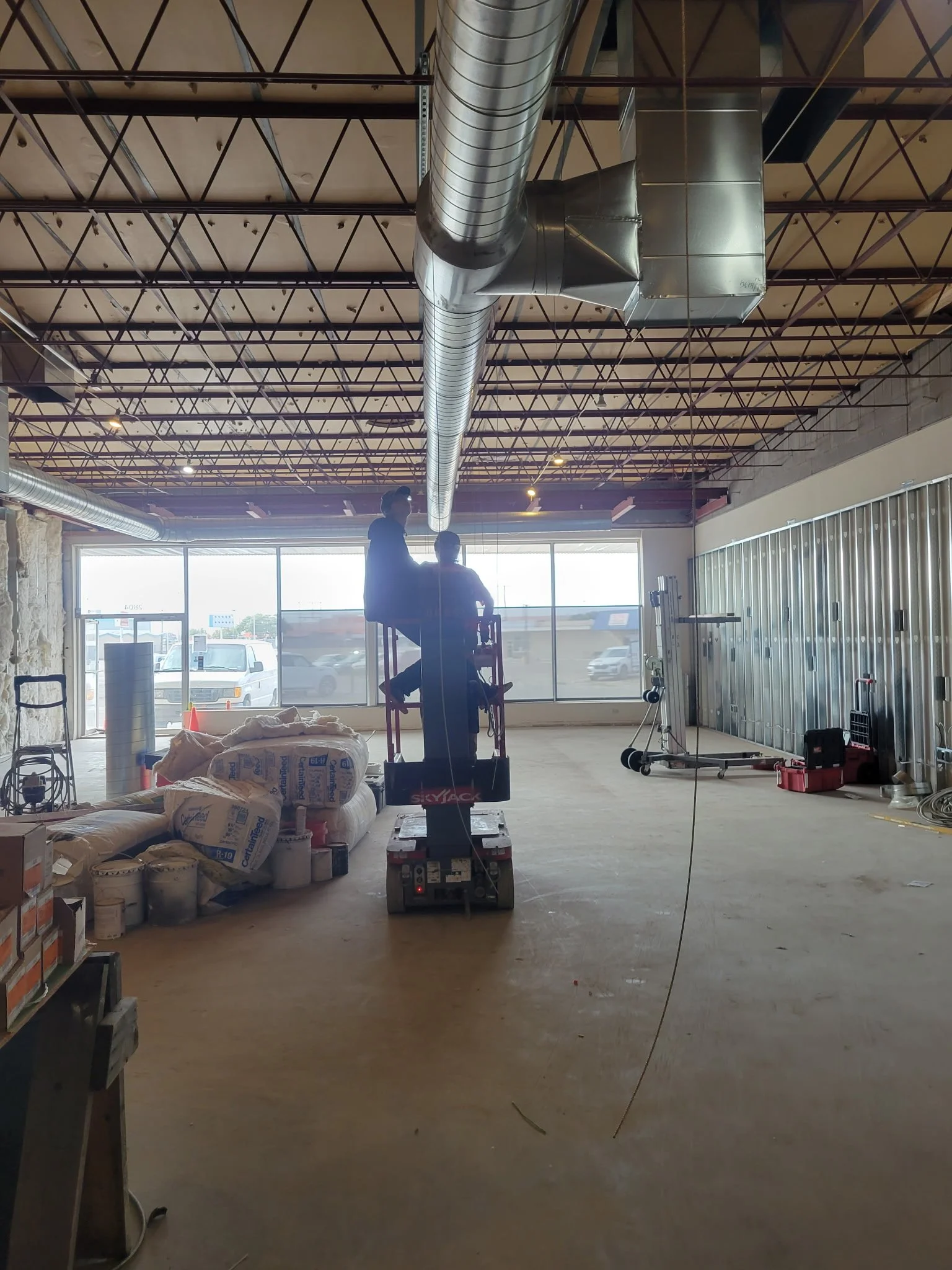 Interior of a building under construction with exposed ductwork, construction materials, and two workers standing on a lift.