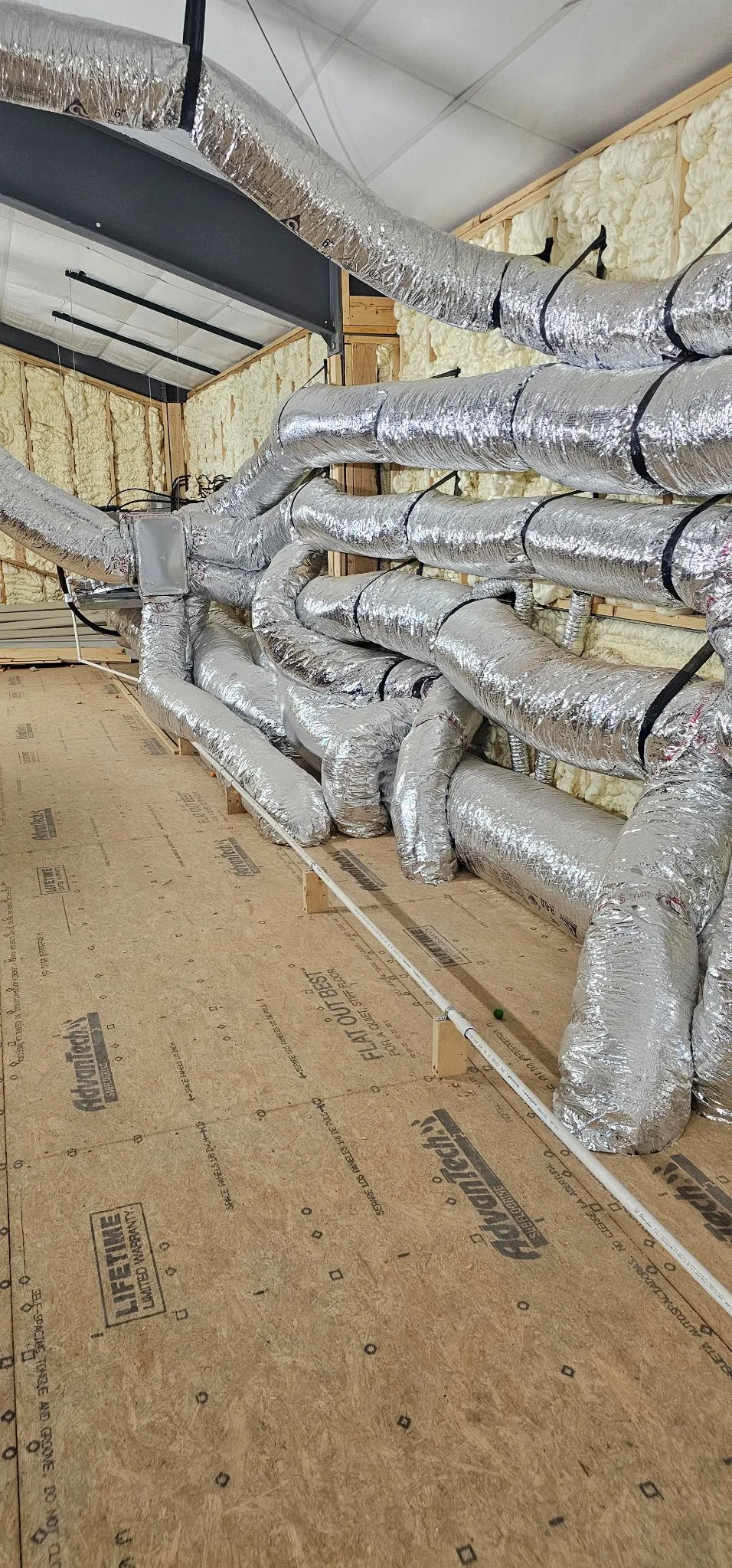 Many different insulated HVAC ducts skillfully placed to be up and out of the way without blocking airflow inside a residential site's attic with insulation material on the walls and a plywood floor.