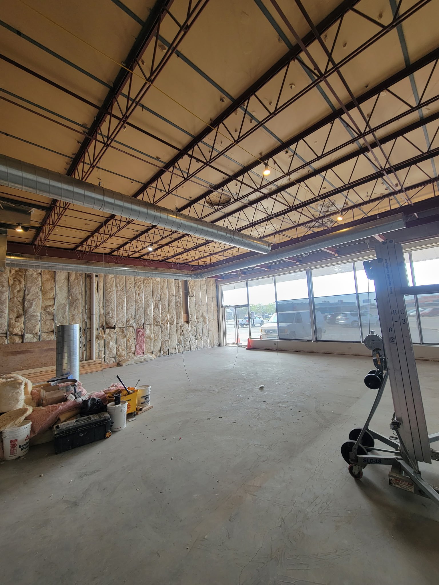Interior of a building under construction with exposed ductwork, insulation, and construction tools, and large windows letting in natural light.