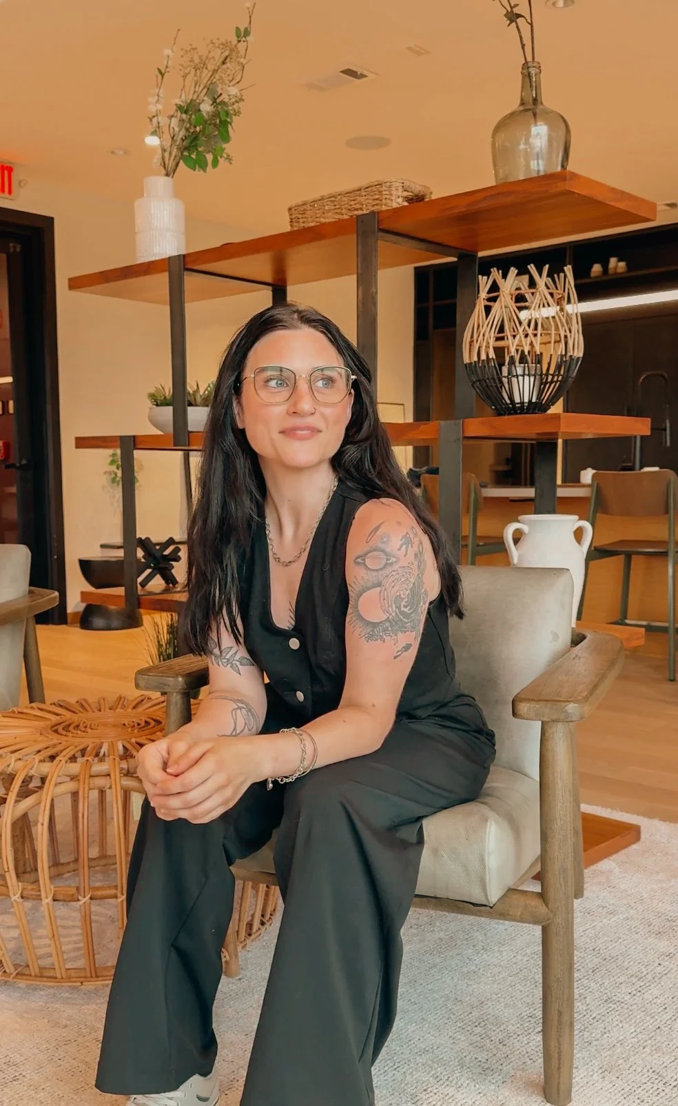 A woman with glasses, long dark hair, and tattoos on her arms, sitting on a wooden chair in a modern, decorated interior space.