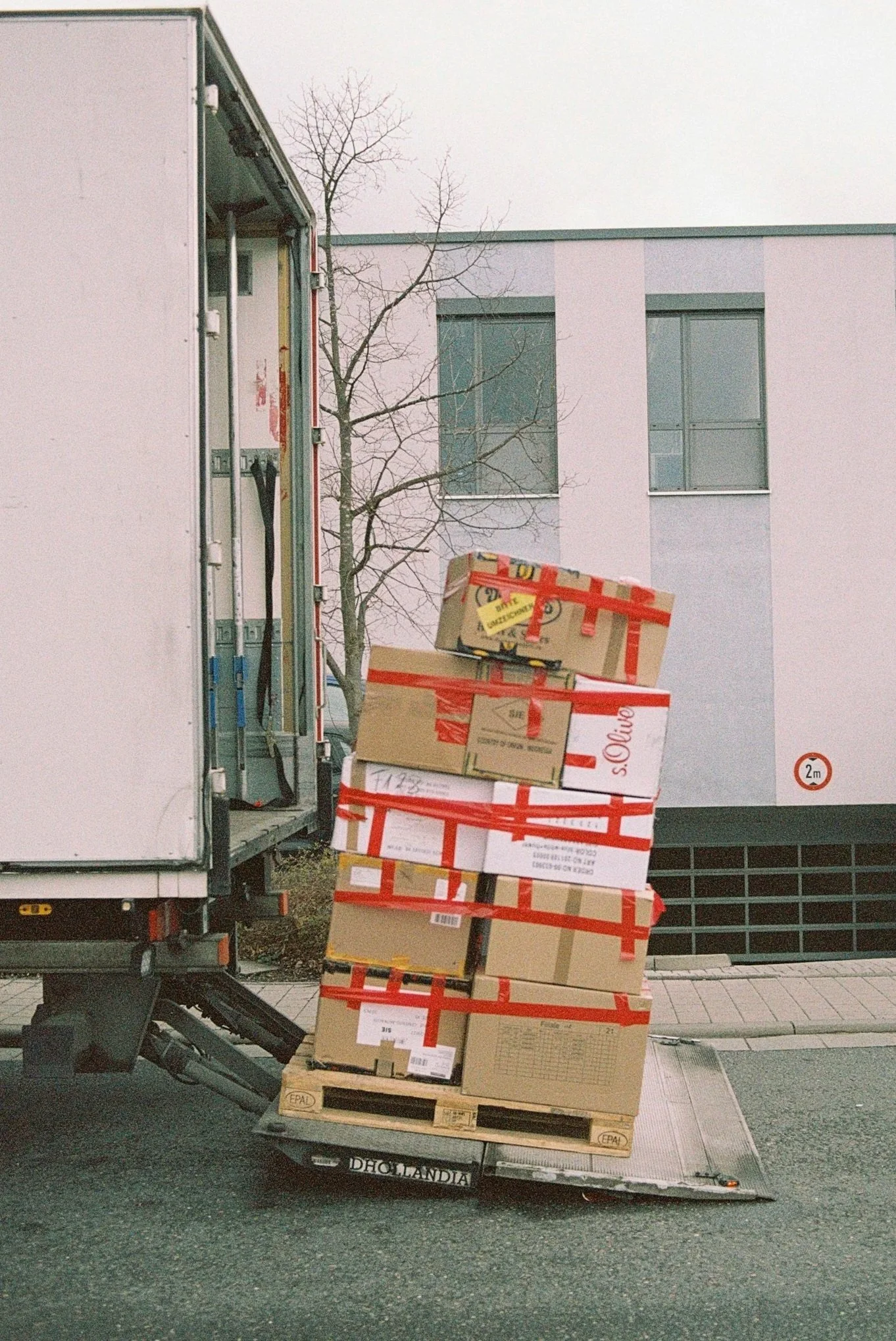 Delivery truck with stacked cardboard boxes on a pallet outside modern building with large windows and a leafless tree in the background.