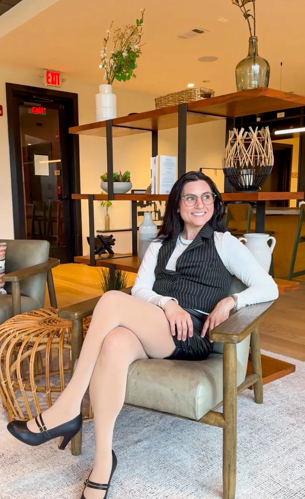 A woman with dark hair and glasses sitting on a beige armchair, smiling, in a modern, well-decorated indoor space with shelves, vases, and plants.