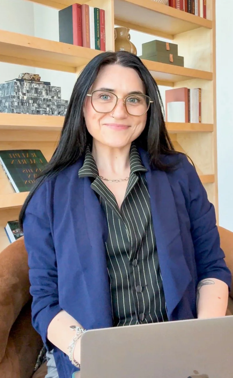 A woman with long dark hair, glasses, and a nose piercing sitting in front of a bookshelf, smiling at the camera, with a laptop in front of her.